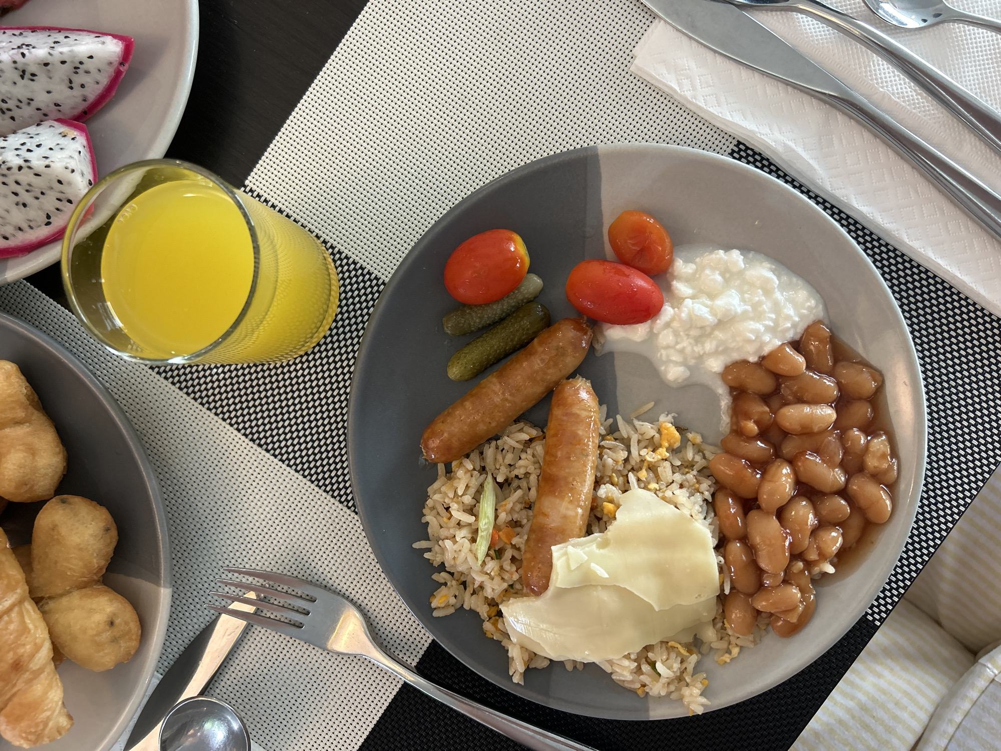 The image depicts a breakfast scene. A grey plate containing fried rice, two sausages, a white sauce (possibly hollandaise), baked beans, cottage cheese, two cherry tomatoes, and two small pickles is centered. Beside the plate are a glass of orange juice and another plate with sliced dragon fruit and pastries. Cutlery is visible on a white napkin. The background consists of a black and white checkered placemat. The scene is located in Rawai, Thailand.