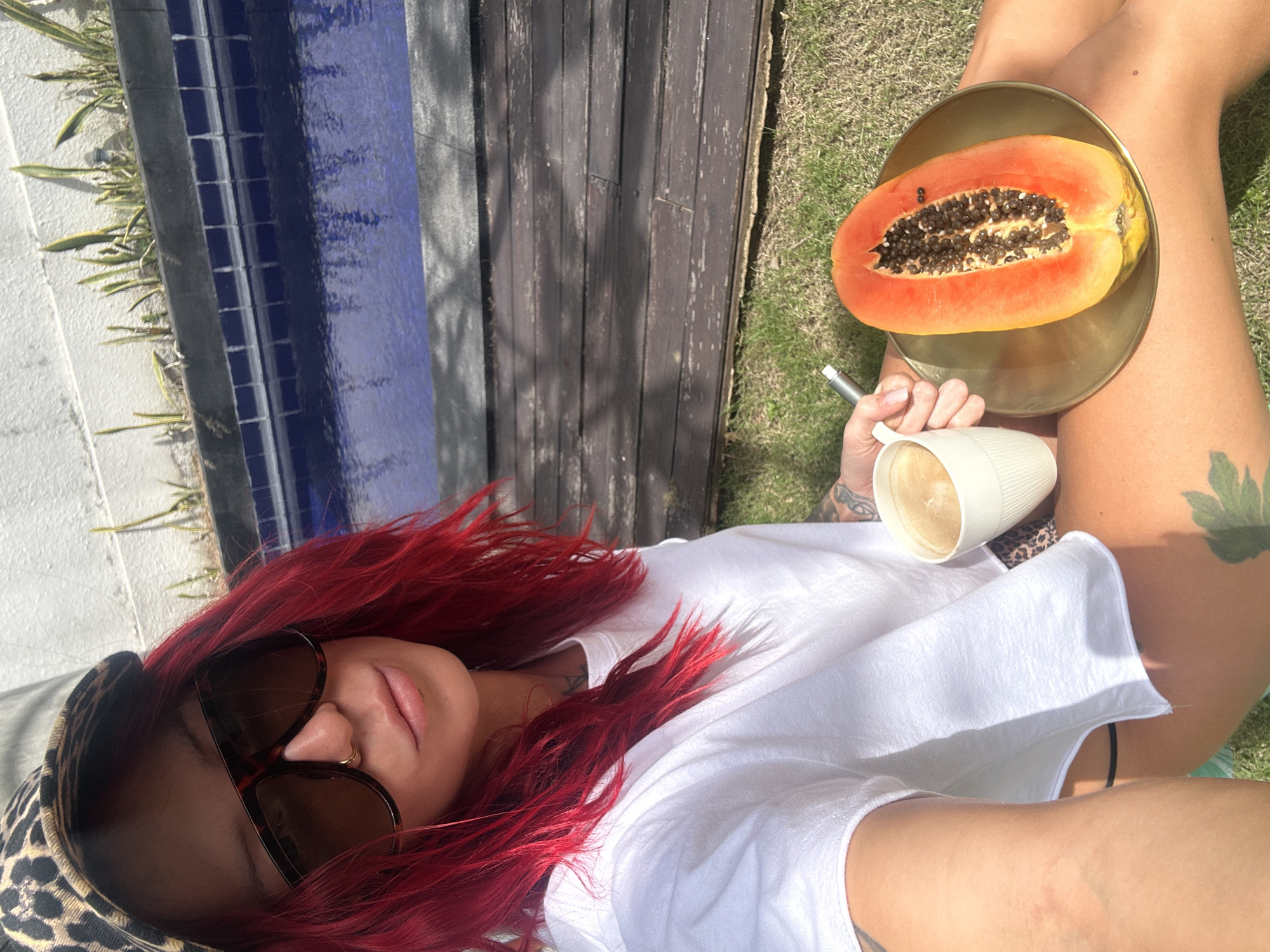 A woman with red hair and sunglasses is lying down, holding a cup of coffee in her left hand, with a halved papaya on a gold plate resting on her legs. The setting appears to be outdoors, possibly in North Kuta, Indonesia, with a blue-tiled wall, wooden paneling, and grass visible in the background. The woman is wearing a white t-shirt and a leopard print cap. The papaya shows a cross-section of its orange flesh and black seeds. Tattoos are visible on her arms and legs. The scene suggests a relaxed, informal setting.