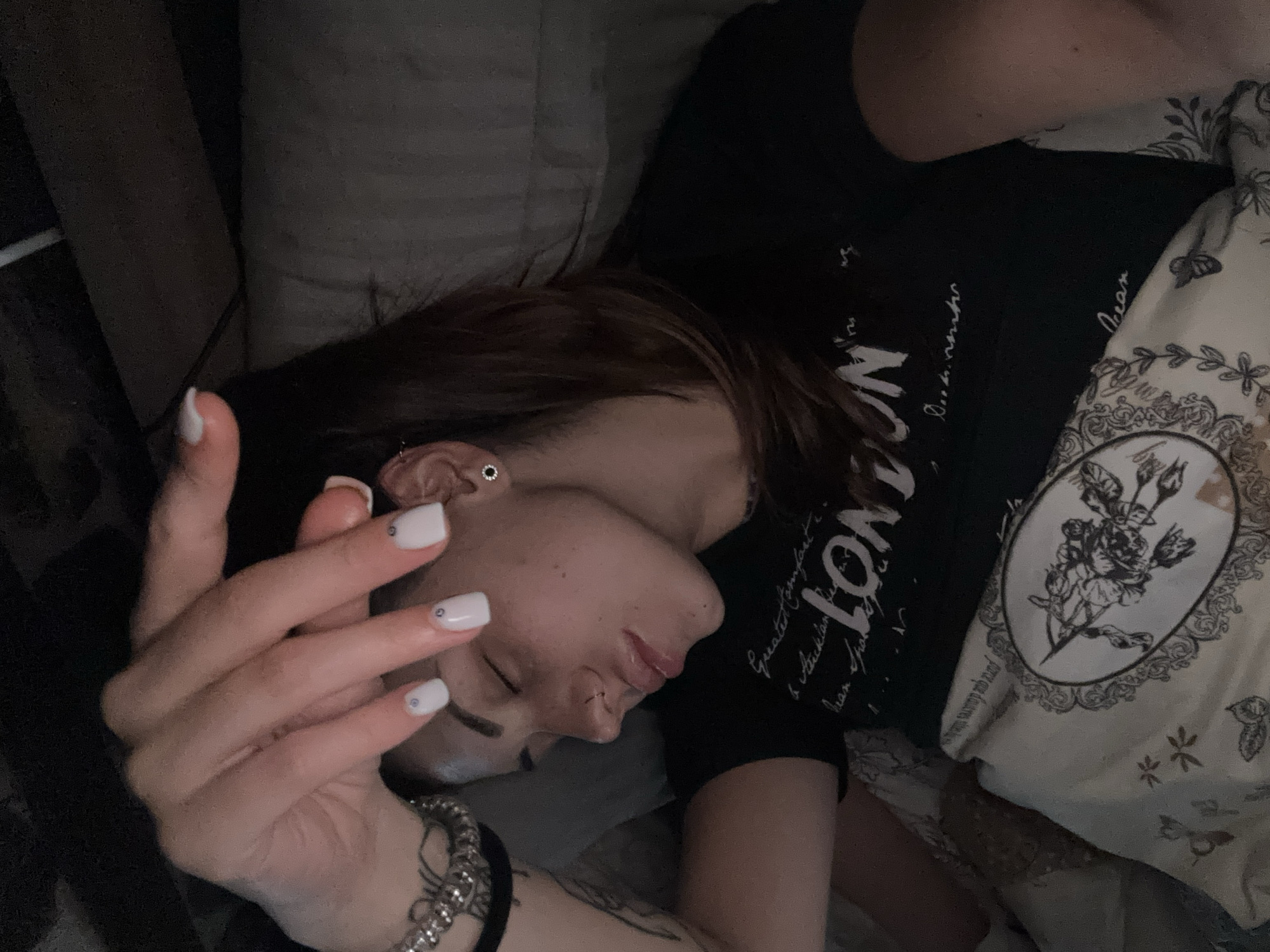 A person is lying in bed with their eyes closed, positioned on their side. The person is wearing a black t-shirt with white lettering, part of which reads "LONDON." The person has short brown hair and is wearing small earrings. Their left hand is raised near their face, with white square-shaped fingernails decorated with small rhinestones. The person is wearing bracelets on their left wrist. Part of a pillow with a rose design is visible. The scene appears to be indoors, presumably in a bedroom. There is no evidence to support the location in the city of Belopoptsi, Bulgaria.