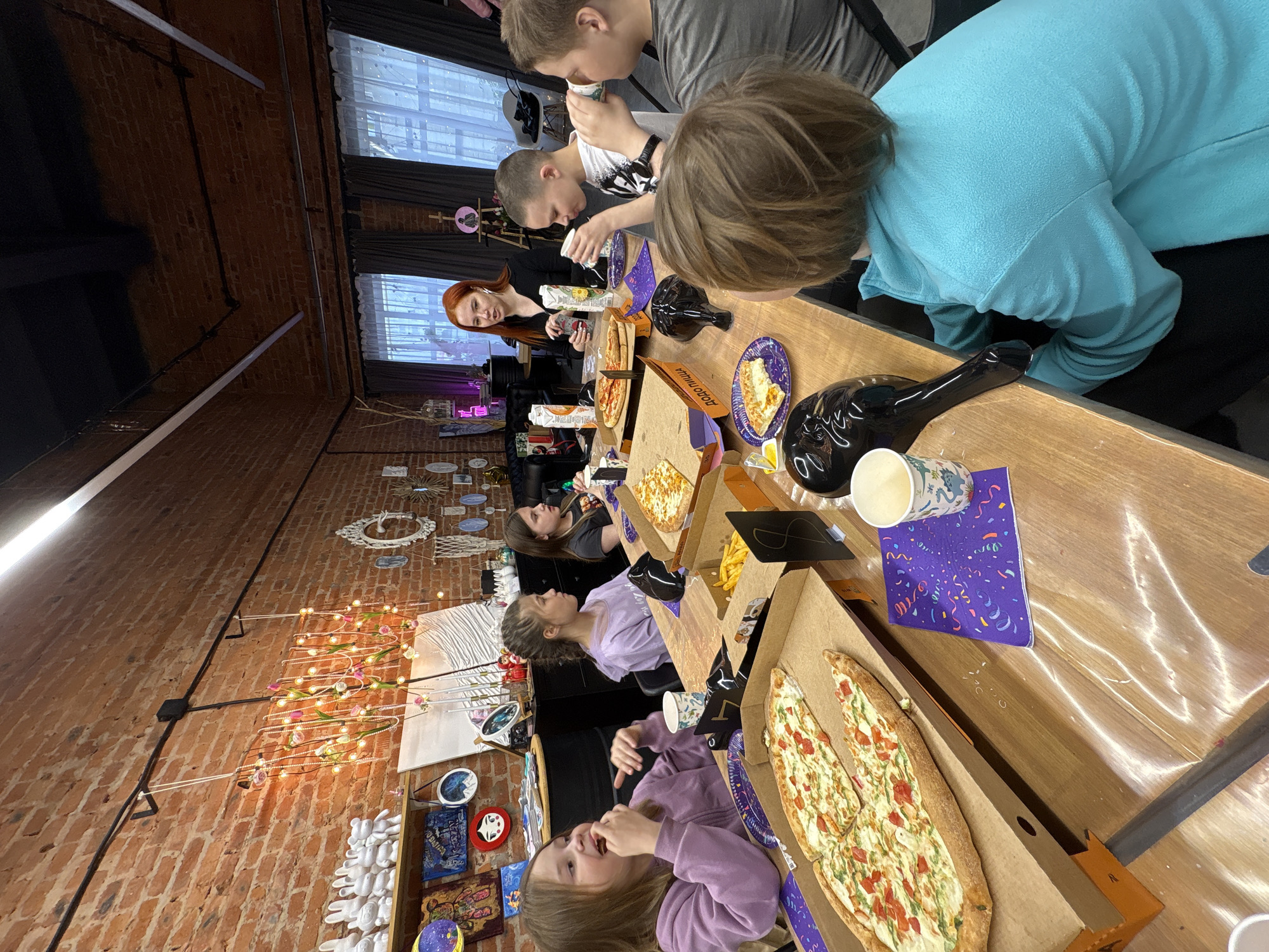 This media file captures a casual indoor gathering, likely a party or social event, set in what appears to be a creative workshop or event space in Chaikovsky, Russia. The scene is dominated by a long, light-colored wooden table around which seven individuals are seated, enjoying food and drinks.
The subjects include a mix of adults and children or teenagers. Towards the far end of the table, a woman with red hair, wearing a black top, is visible. Along the table, several young people are seated, including two boys at the head of the table, one in a grey t-shirt and another in a white t-shirt. Further down, three girls or young women are present, all wearing shades of purple or lavender tops. One girl looks towards the camera with a slightly open mouth, possibly mid-chew, indicating a moment of enjoyment. Another person in a blue fleece jacket has their back to the camera.
The main activity is eating and socializing. The table is filled with food, primarily several open pizza boxes containing elongated, flatbread-style pizzas topped with cheese, diced tomatoes, and green herbs. Some pizza slices are served on individual purple paper plates decorated with a confetti pattern. A container of french fries is also visible. People are drinking from patterned paper cups, and a distinctive black, bottle-shaped ceramic or glass object is also on the table.
Notable details include the setting's aesthetic: exposed brick walls suggest an industrial or rustic feel, complemented by large windows with sheer white curtains that let in natural light, indicating it's daytime. Decorative elements on the brick wall include string lights adorned with flowers, various circular artworks, and white ceramic hand shapes, contributing to a creative or artistic atmosphere. Visible text on one of the pizza boxes clearly spells "ДОДО ПИЦЦА" (Dodo Pizza), identifying the food provider. A purple napkin also has the word "MY" printed on it. The overall mood appears relaxed and cheerful.