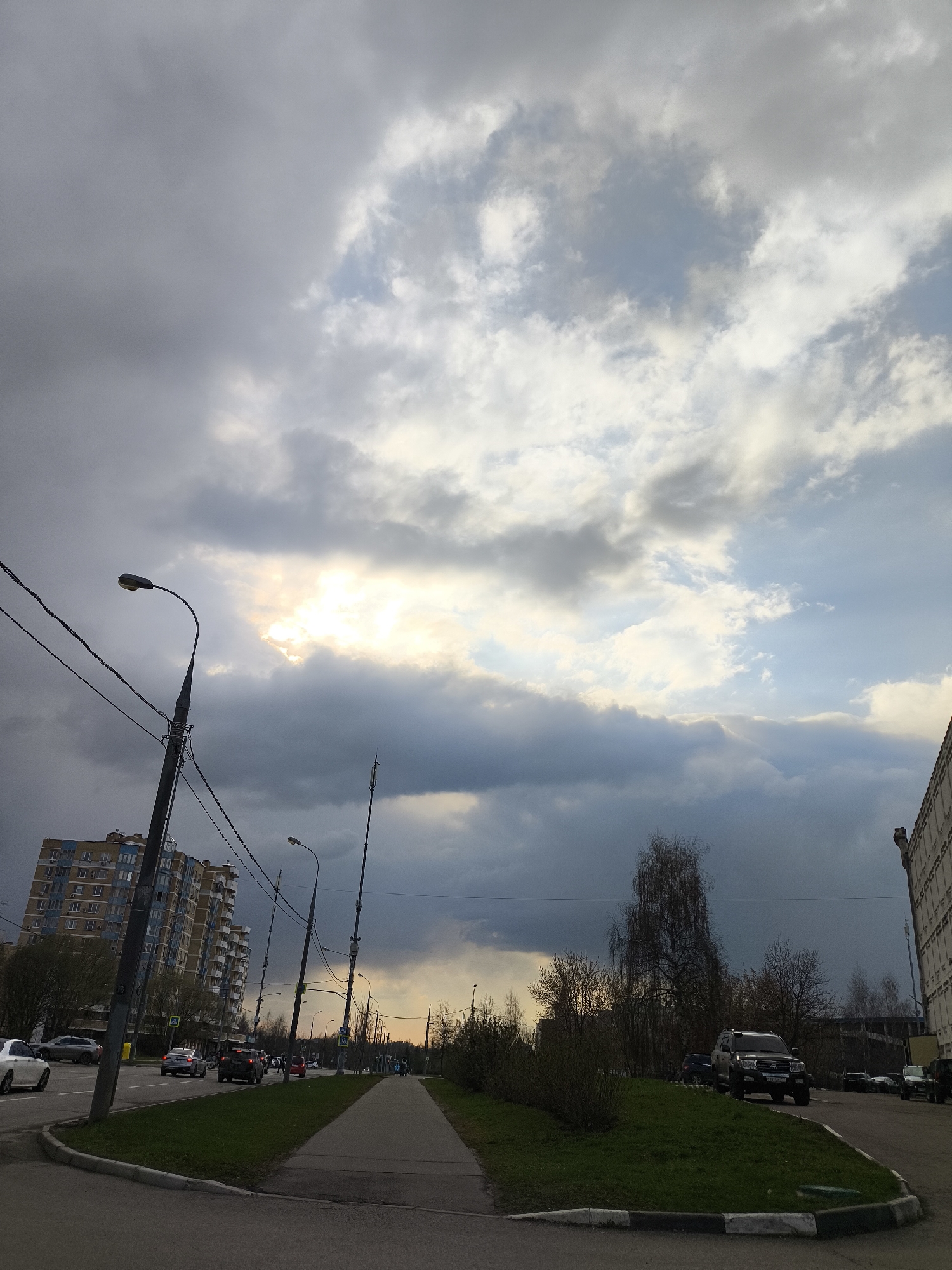 The image depicts a street scene in Moscow, Russia, under an overcast sky with patches of sunlight breaking through the clouds. A multi-story apartment building is visible on the left, while a taller, plain building stands on the right. A sidewalk runs along the edge of the street, bordered by patches of green grass and low bushes. Cars are seen driving on the road in both directions. Utility poles line the street, with power lines crisscrossing the sky. In the distance, trees and the horizon line suggest an urban or suburban setting. The overall impression is that of a typical city street under cloudy weather.