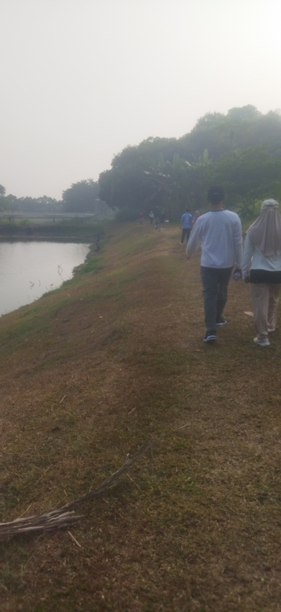 The image depicts a hazy outdoor scene, likely in Depok, Indonesia, possibly during the morning or late afternoon. In the foreground, a dry, grassy path slopes upwards, with scattered dry twigs and leaves on the ground. To the left, a body of water, possibly a lake or pond, reflects the muted sky. In the mid-ground, several individuals are walking along the path, their backs to the camera. Two figures in the front are more clearly visible: one wearing a white long-sleeved shirt and dark pants, and another in a light-colored hijab and light-colored top with dark pants. Other figures are further up the path, partially obscured by distance and haze. The background is dominated by dense, dark green foliage, suggesting trees and vegetation on a hill, with the hazy sky above. A bridge or elevated structure is faintly visible in the far left background across the water. The overall atmosphere is calm and appears to be a recreational setting.