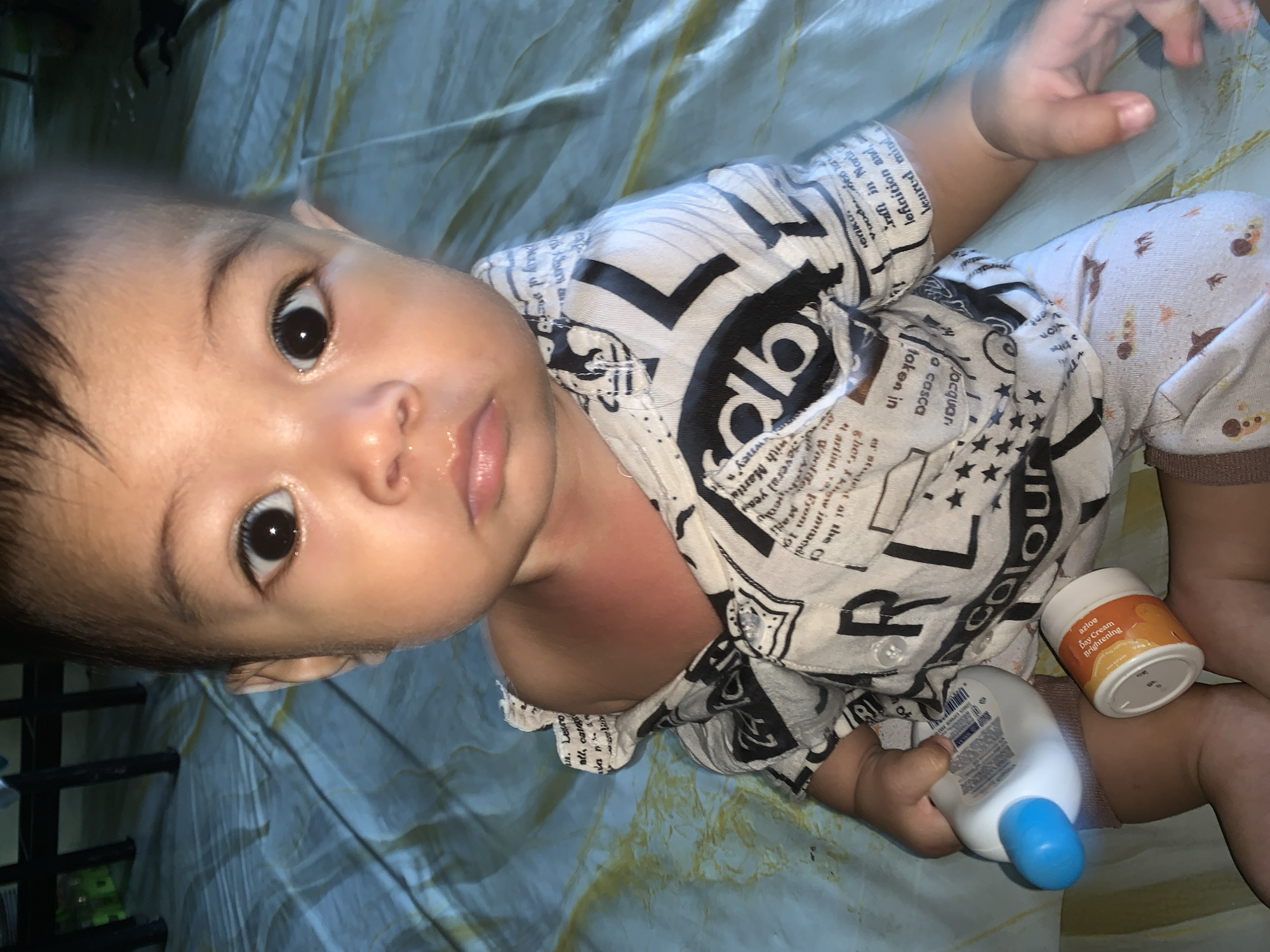 A close-up shot of a baby lying on a bed sheet with a bottle and cream on their right side. The baby has dark hair and eyes, and is wearing a black and white patterned shirt and patterned shorts. The cream is a white jar with an orange label that reads "azloe Day Cream Brightening". The bottle has a blue cap. The baby is looking up at the camera with a slightly open mouth.
The location is assumed to be a residence or care facility within Sungai Meriam, Indonesia, based on the assumption that the picture taker is familiar with the area. There is no location information visible within the image.
