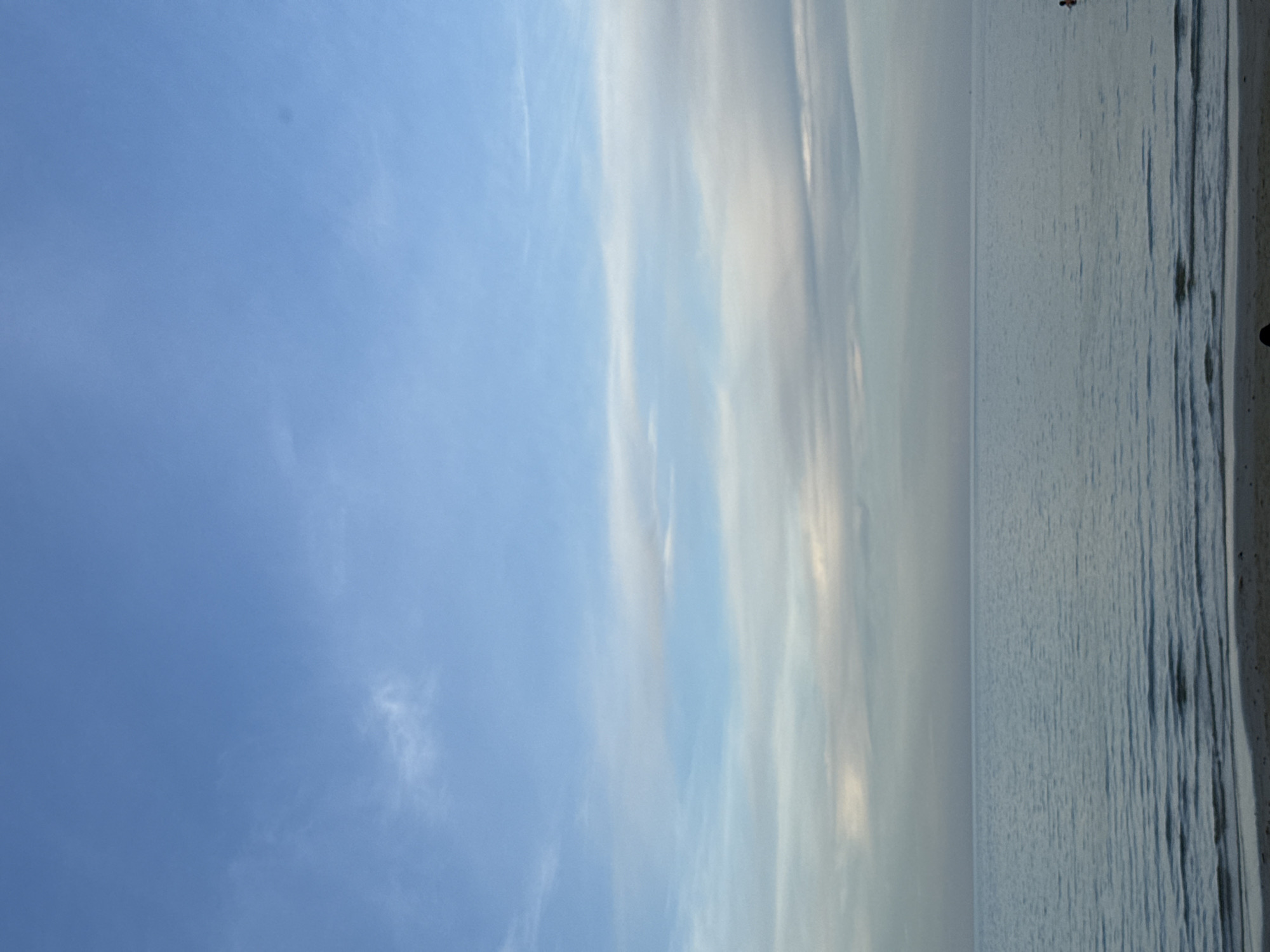 The image depicts a serene seascape under a partly cloudy sky. The horizon is clearly defined, separating the calm, blue water from the sky. The sky is a gradient of light blue with wispy white clouds scattered across it, becoming denser and grayer towards the horizon. The water exhibits gentle ripples, reflecting the sky's muted colors. A strip of sandy beach is visible in the foreground, with subtle wave patterns lapping at its edge. In the distance, on the right side of the frame, a small dark silhouette of a person is visible in the water, appearing to be partially submerged. The overall impression is one of tranquility and natural beauty.