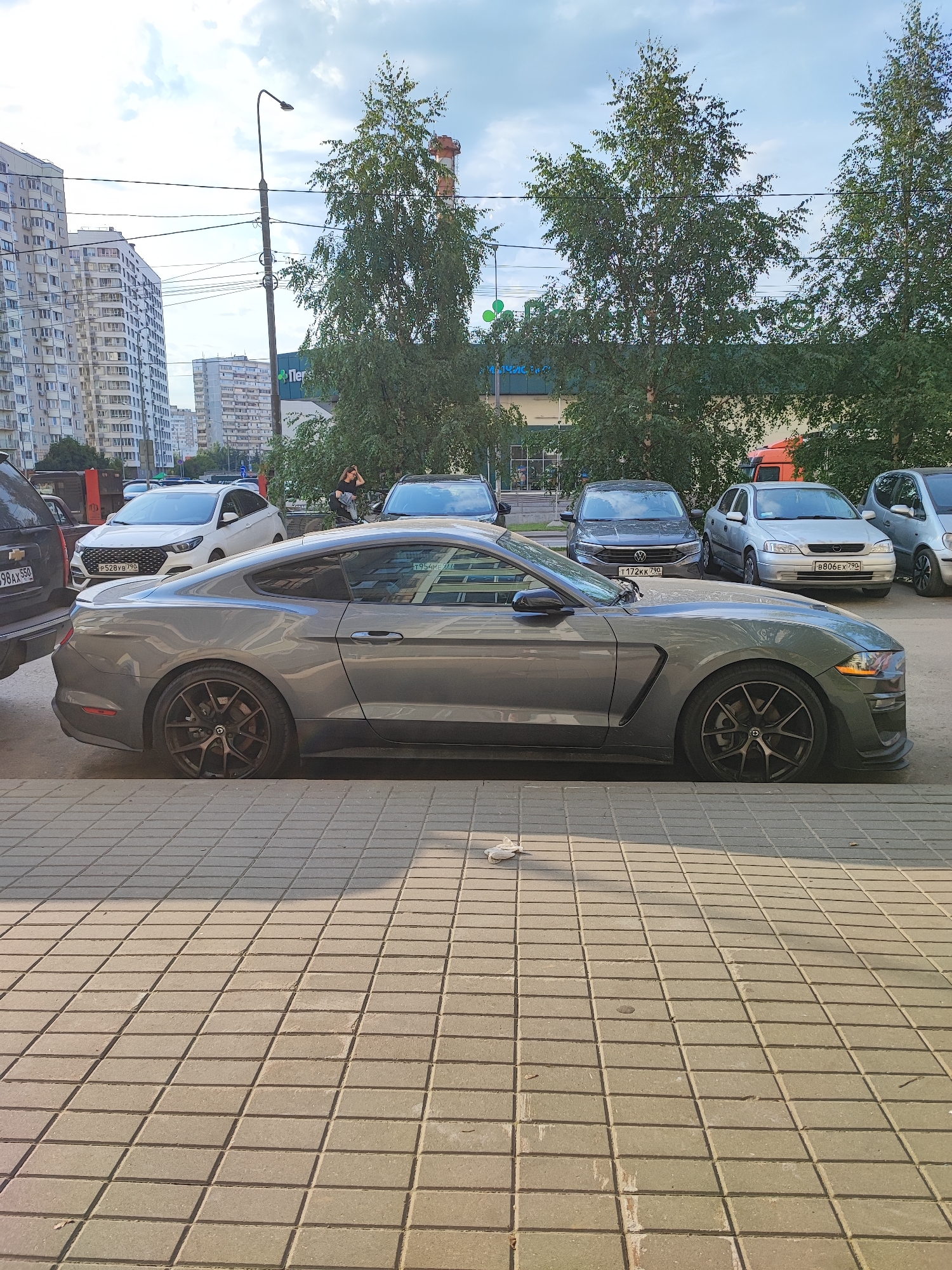 The image captures a street scene in Vidnoye, Russia, likely during daytime under a cloudy sky. A grey Ford Mustang, a two-door coupe with custom wheels, is parked along a sidewalk paved with light-colored square tiles. The car is positioned parallel to the curb, facing right. In the background, several other vehicles are visible: a white sedan, a dark sedan with a VW logo, a silver sedan, and a silver hatchback. Behind the cars, a row of trees with green foliage stands in front of a building with a green awning, possibly a commercial establishment. Further in the distance, apartment buildings with numerous windows dominate the skyline. A lamppost with wires stretches across the scene. A person, wearing dark clothing, is visible standing near the trees, with their hand touching their head. There is also a white object, possibly trash, on the tiled sidewalk in the foreground. The overall atmosphere suggests an urban environment.