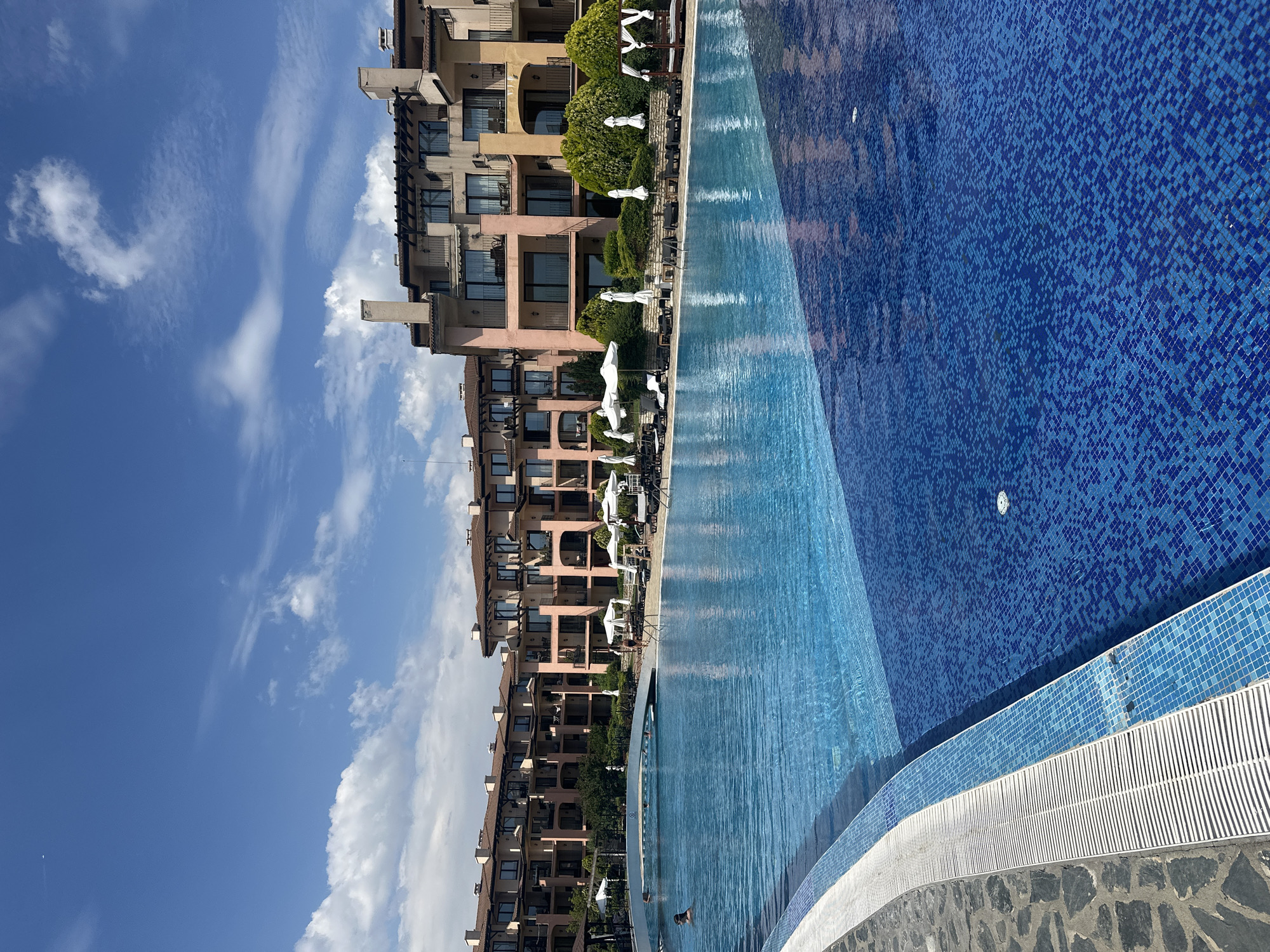 This media file features a picturesque scene of a resort with a large, inviting swimming pool. The pool itself is a dominant element, with its vibrant blue mosaic tiles creating a shimmering surface that reflects the bright sky. The water appears calm and clear, with gentle ripples suggesting it is being used.
In the background, a multi-story building with a warm, sandy-colored facade stands tall. The architecture features numerous balconies, arched windows, and rooflines that suggest a Mediterranean or tropical resort. Lush green trees and shrubs are neatly arranged along the building's base and around the pool area, adding a touch of natural beauty to the scene. White umbrellas are open, providing shade for lounge areas beside the pool, hinting at a relaxing atmosphere.
The sky above is a brilliant blue, dotted with fluffy white clouds that indicate fair weather. The sunlight is strong, casting clear shadows and illuminating the scene, suggesting it is daytime, likely mid-morning or afternoon. There are a few people visible in the pool, appearing to be enjoying a swim. Their presence adds a sense of life and activity to the otherwise serene setting.
The overall impression is one of a luxurious and tranquil vacation destination, perfect for relaxation and enjoyment. The composition, with the pool in the foreground and the building and sky in the background, creates a sense of depth and immersion. No visible text is present in the image.