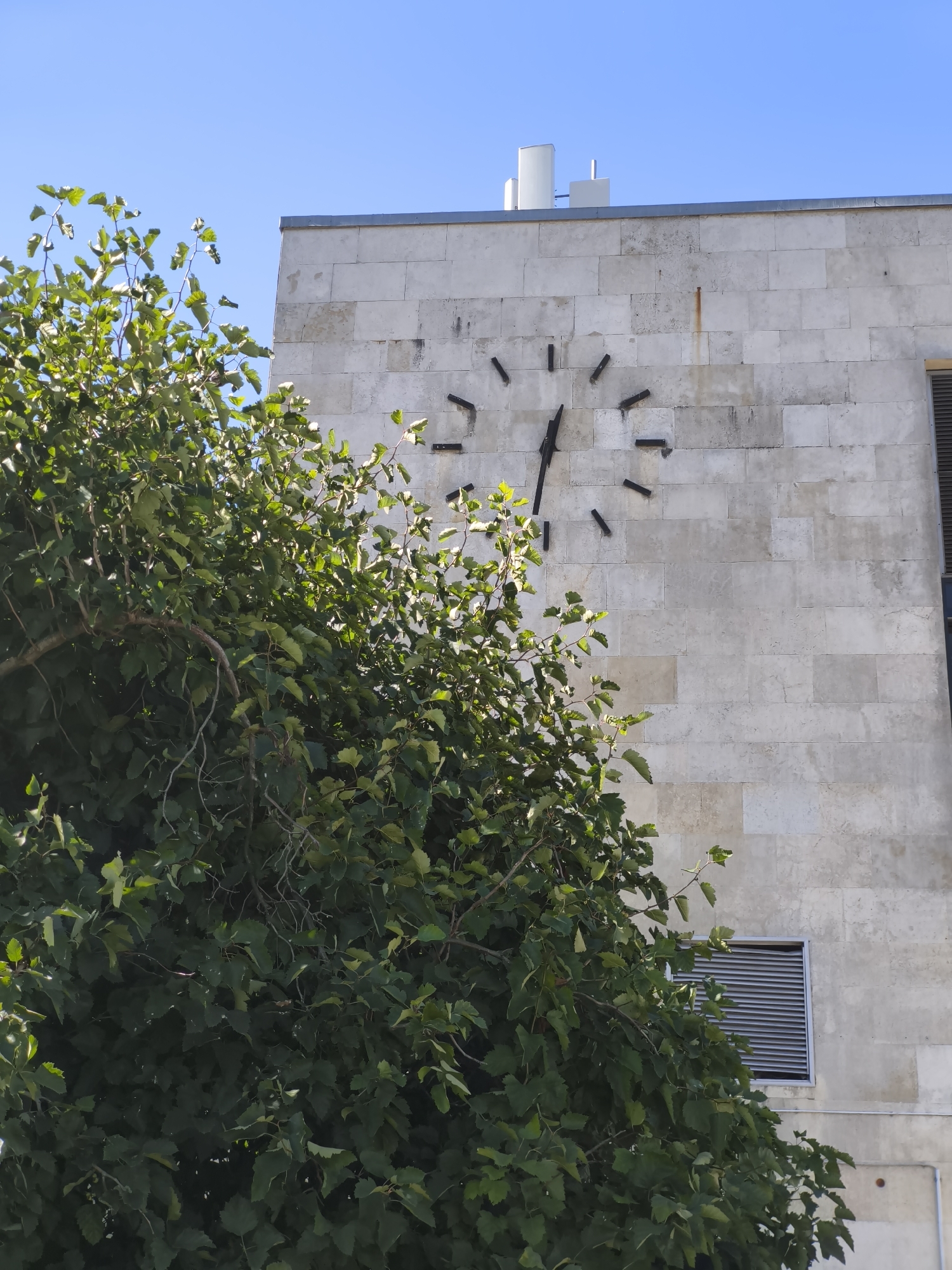 A large, outdoor clock is mounted on a light-colored, stone-clad building. The clock has no numbers, only black, rectangular markers for the hours, and black hands indicating the time. The hour hand points to approximately 10:30, while the minute hand points to approximately 3:00. In the foreground, a large tree with abundant green leaves partially obscures the view of the clock and building. The sky above is a clear, bright blue. The building appears to be modern, with a minimalist design. The presence of louvred vents on the side of the building and what appear to be communication antennas on the roof suggest an institutional or commercial building. This building is the Kumu Art Museum, located in Tallinn, Estonia.