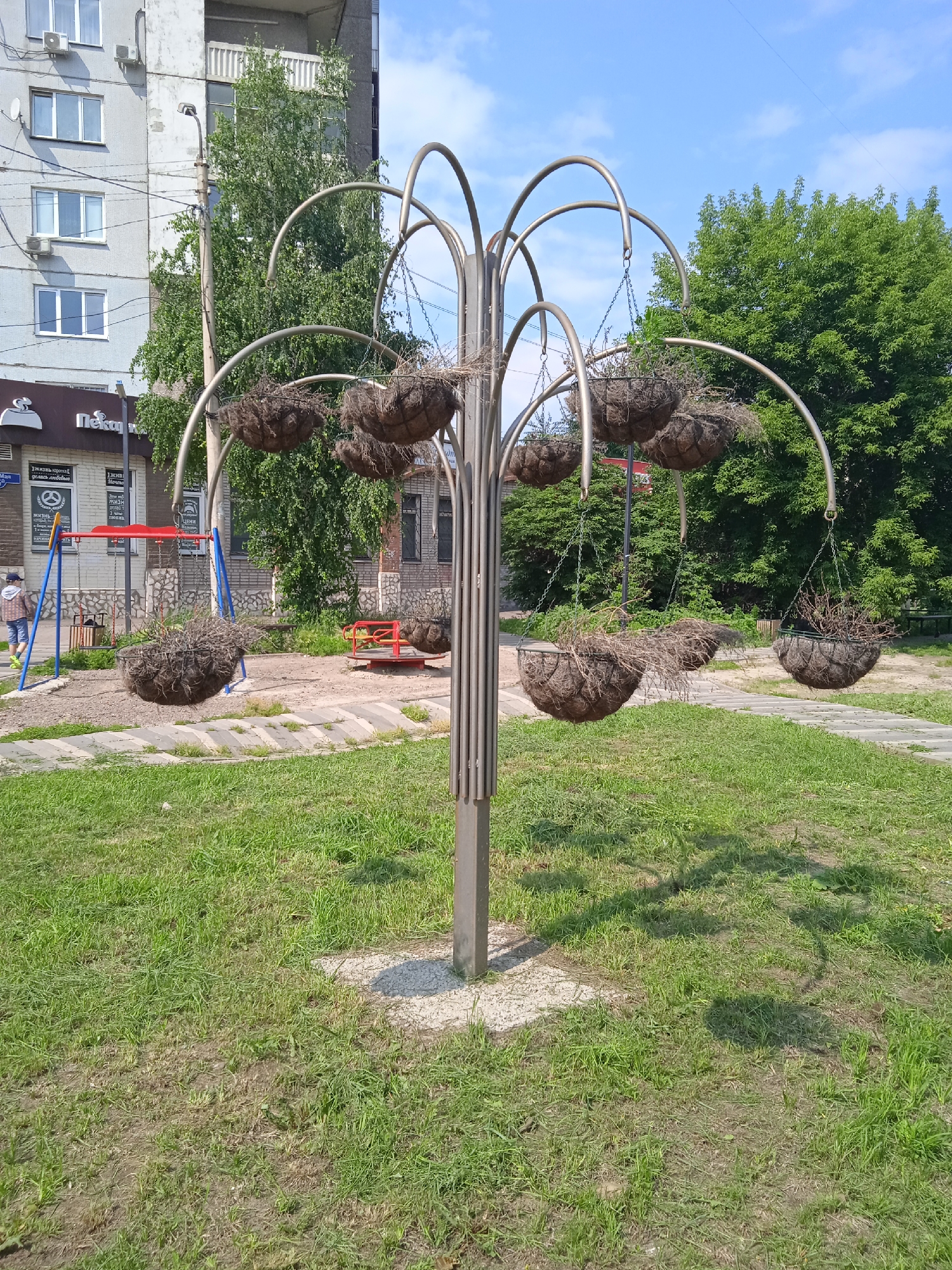 The image shows an outdoor metal structure designed to hold hanging planters. The structure resembles a stylized tree with multiple curved metal arms supporting round, coir-lined planters. Most of the planters are empty or contain dry plant debris. The structure stands on a concrete base surrounded by mowed grass. In the background, there is a children's playground with a swing set and a merry-go-round, suggesting a park or residential courtyard. Behind the playground are buildings, including a multi-story apartment building and a smaller commercial building with signage. Lush green trees and a bright blue sky with scattered clouds complete the scene. The location is identified as Krasnoyarsk, Russia.