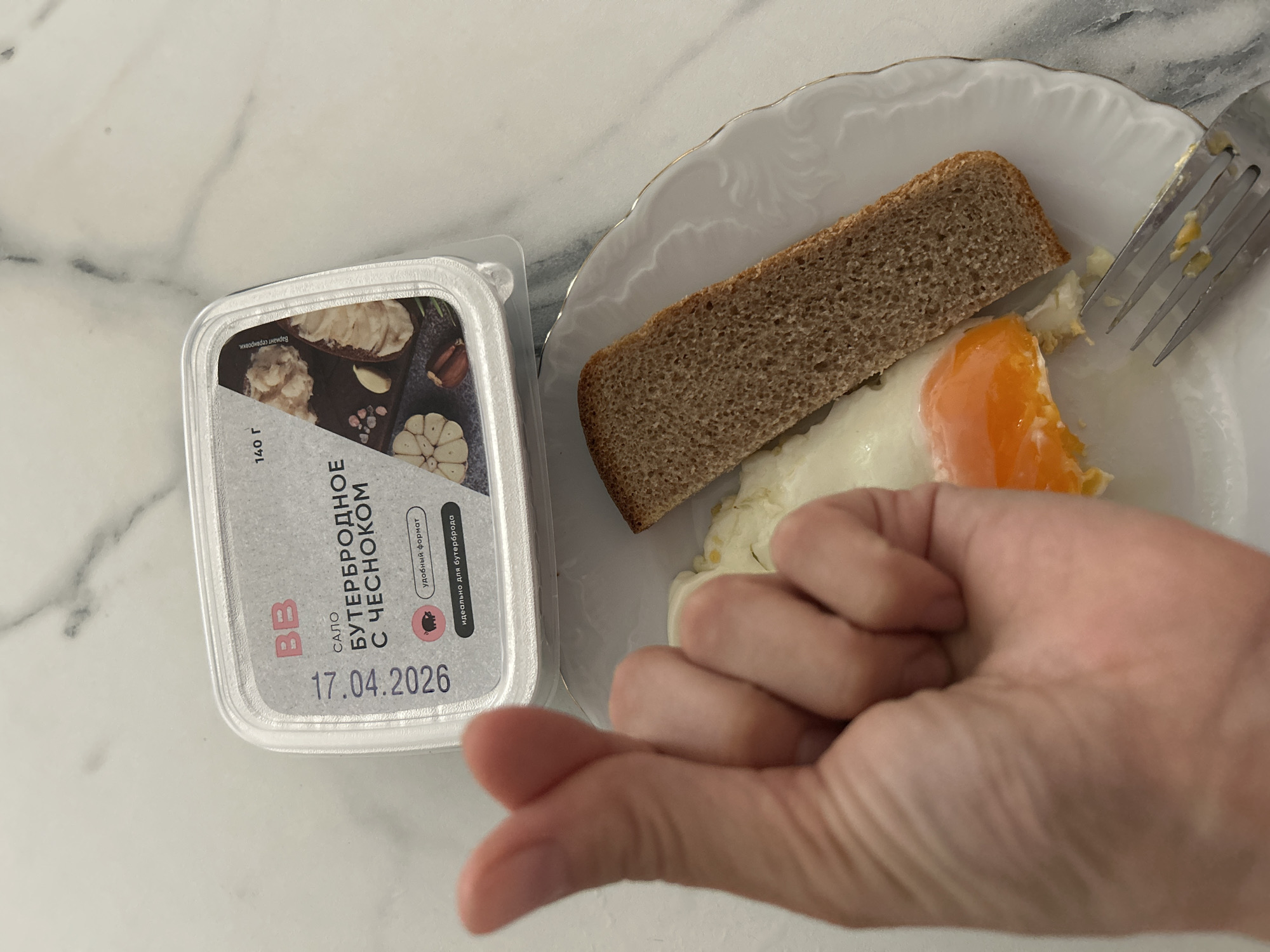 The image displays a close-up, overhead view of a meal arranged on a light-colored, possibly marble or granite, surface. The main subjects include a white plate with a decorative scalloped edge and gold rim, holding a slice of dark bread and a fried egg with a bright orange, partially broken yolk. A silver-colored fork rests on the edge of the plate.
Adjacent to the plate is a plastic container of food spread, featuring a label predominantly in Russian. Visible text on the label includes "BB" (likely a brand name), "САЛО БУТЕРБРОДНОЕ С ЧЕСНОКОМ" (Salo sandwich spread with garlic), "140 г" (140 grams), "удобный формат" (convenient format), and "идеально для бутерброда" (ideal for a sandwich). A prominent date, "17.04.2026", is also visible, indicating a best-before or expiration date. The label's design includes an image of garlic cloves and a slice of bread with the spread. A person's hand, with fingers curled, is partially visible in the bottom right corner, suggesting interaction with the setting.
The scene depicts an indoor setting, likely a kitchen or dining area in Moscow, Russia, given the Cyrillic text and the nature of the "salo" product. The overall impression is of a simple meal or breakfast being prepared or consumed. No specific time of day or weather conditions are discernible.