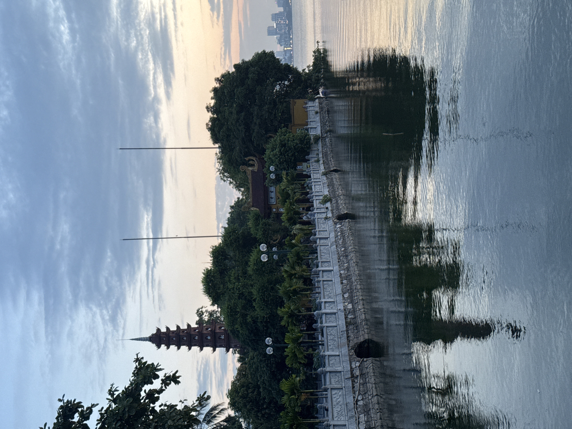 This is a vertical, eye-level shot of Tran Quoc Pagoda, located on West Lake in Hanoi, Vietnam, at sunset. The pagoda, a red, multi-tiered structure with a spire, is visible above a dense canopy of green trees. In front of the trees, a long, ornate stone wall runs along the water's edge, punctuated by streetlights and palm trees. The water reflects the sky, trees, and pagoda. In the distance, a cityscape with modern buildings is visible, bathed in the soft light of the setting sun. The sky is cloudy with streaks of orange and pink.