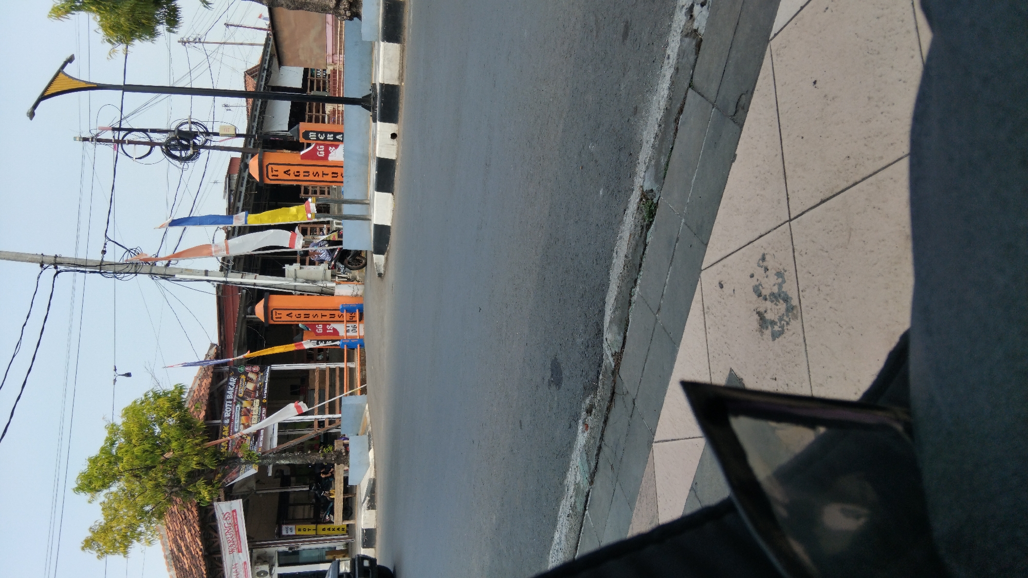 The image depicts a street scene in Batang, Indonesia, likely on or around August 17th, given the prominent "17 AGUSTUS" signage on two orange pillars. The scene appears to be in front of a business, possibly a food stall or restaurant, as indicated by the signs "ROTI BAKAR" (grilled bread) and "WARUNG SS". Several flags, including red and white Indonesian flags, are displayed, suggesting a national holiday celebration. A few people are visible, one on a motorbike and others possibly inside the establishment. The street itself is paved, with a sidewalk made of checkered tiles. The environment is urban, with utility poles and electrical wires overhead.