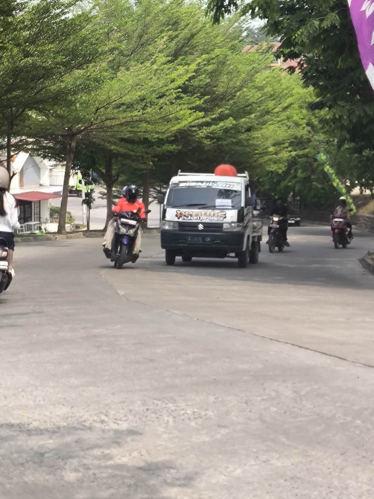 The image depicts a street scene in Cilegon, Indonesia, with a focus on vehicular traffic. A white Suzuki Carry pickup truck with a red circular object on its roof is the most prominent vehicle in the foreground, traveling on a paved road. The truck has various decals on its doors and windshield. To the left of the truck, a person wearing an orange shirt and a helmet is riding a dark-colored motorcycle. Further to the left, a person in a white top and helmet is on a scooter, facing away from the camera. In the background, other motorcycles and vehicles are visible, along with lush green trees lining the street and some buildings. The overall impression is of a typical day on a city street with moderate traffic.