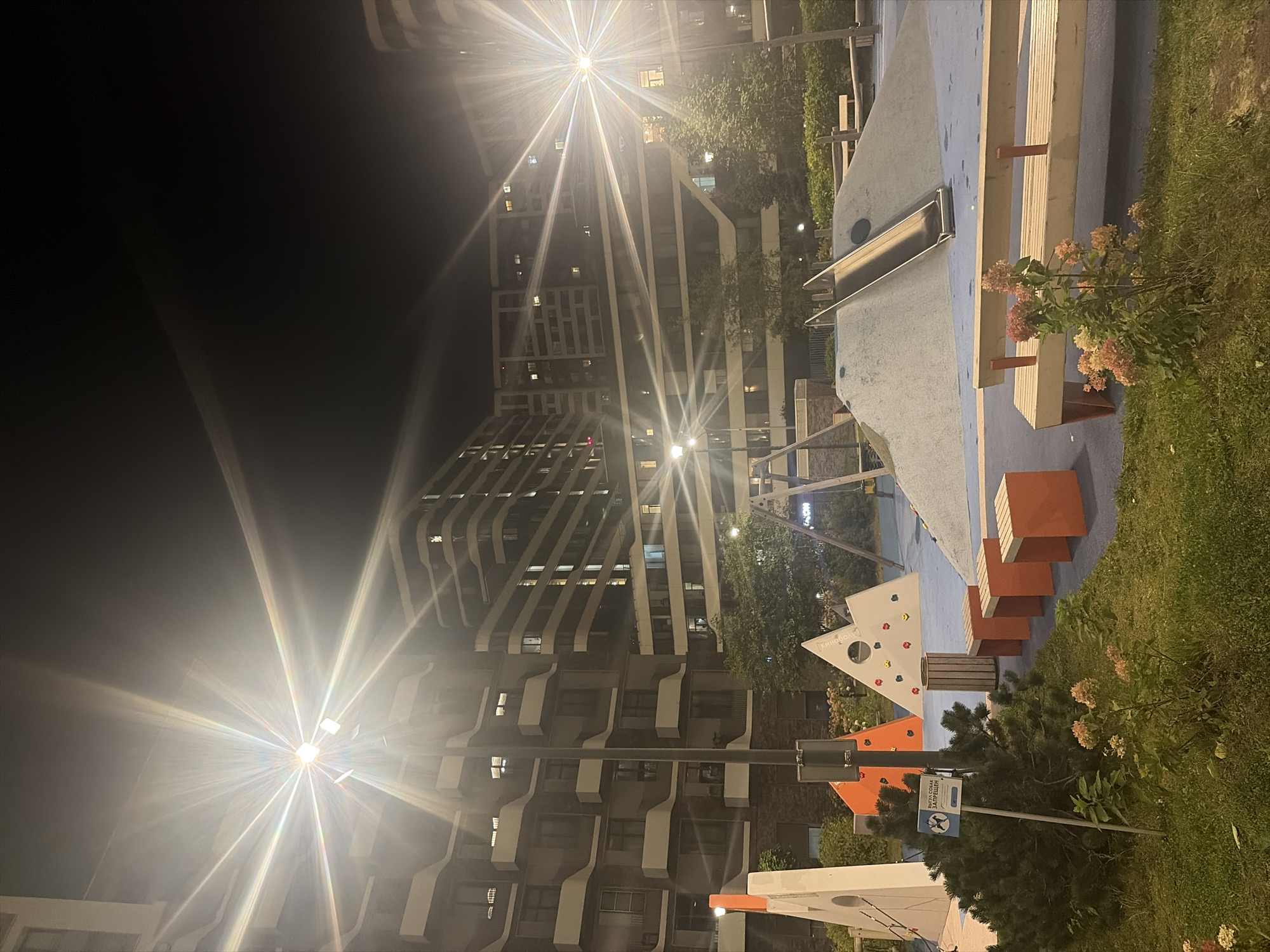 The image shows a nighttime view of a modern residential complex in Moscow, Russia. In the foreground, there is a children's playground with various structures, including slides, climbing walls designed as geometric shapes, and seating benches. The playground is illuminated by bright streetlights, which create lens flares and add a dramatic effect to the scene. The surrounding area is a grassy lawn with some bushes and flowering plants. The residential buildings are tall and modern, with many windows lit from within, indicating that people are inside. The overall atmosphere is calm and serene, despite the harsh lighting.