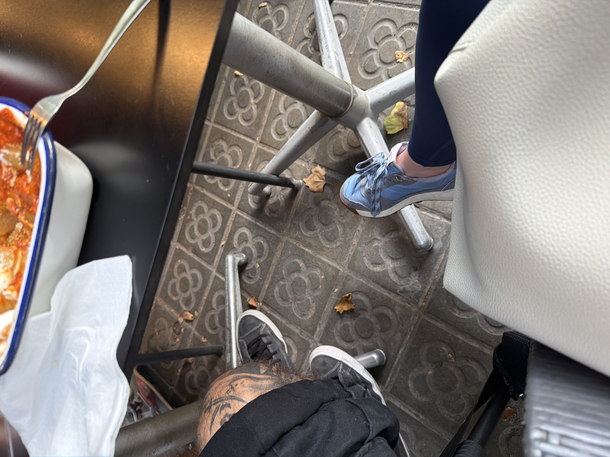 The media file depicts an outdoor scene at what appears to be a cafe or restaurant, possibly in Barcelona given the characteristic patterned tiles on the ground. The image is taken from a high-angle perspective, looking down at the lower legs and feet of two individuals seated at a table.
One person is wearing blue athletic shoes with white laces and a light pink trim around the ankle. They are also wearing dark blue leggings or trousers. Their foot is resting near the metal base of a table or chair.
The other person has black canvas sneakers with white laces and white soles. Their feet are positioned closer to the foreground, and they are wearing black trousers or jeans. This individual also has visible tattoos on their leg, featuring intricate designs with shading.
In the background, a white chair or stool with a textured surface is partially visible. A portion of a dish containing food, likely a pasta dish with a red sauce, is seen in the upper left corner, with a fork resting in it. A white napkin is also present on the table. The ground is paved with grey square tiles, each featuring a distinct, repeating floral or clover-like pattern, a common design found in Barcelona. A few dry fallen leaves are scattered on the ground, suggesting it might be autumn. The lighting suggests it is daytime, with diffused natural light. The overall atmosphere appears relaxed, indicative of a casual meal or break.