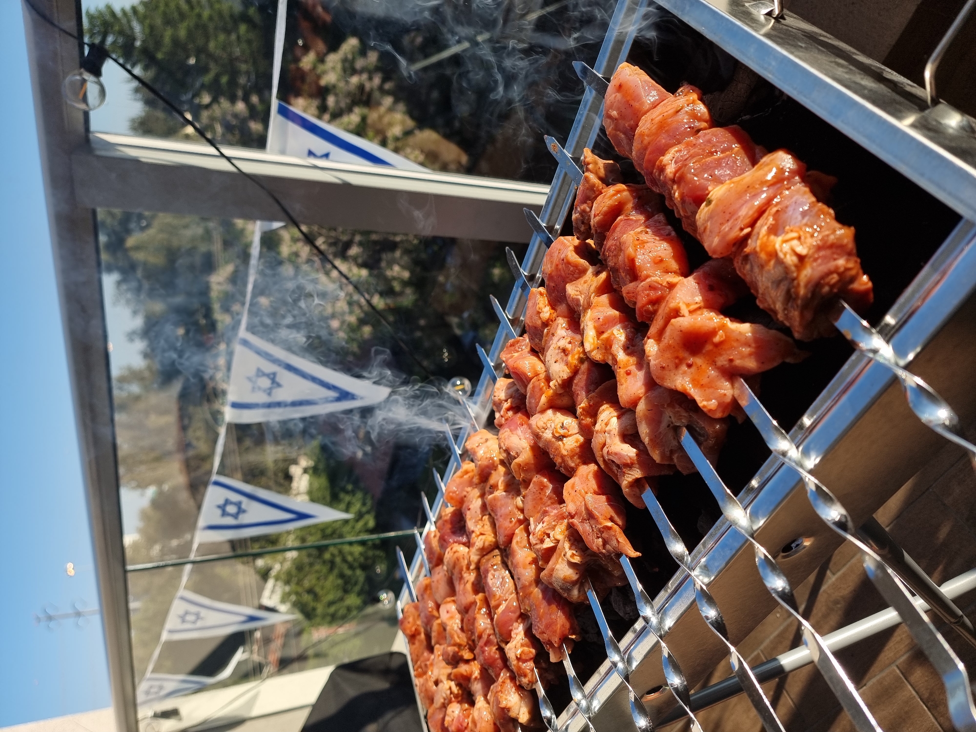 This vibrant image captures an outdoor barbecue scene, likely set in Ramat Gan, Israel, during clear daytime hours. The primary focus is a metal grill, heavily laden with numerous skewers of marinated meat, possibly chicken or pork, charring over hot coals. The meat chunks, coated in a reddish-orange marinade, glisten under the sun, with fine wisps of smoke rising from the grill, hinting at the cooking process. The metallic skewers feature distinct twisted handles.
In the midground, behind what appears to be a glass barrier or windowpane, a string of small, triangular Israeli flags is visible. Each flag proudly displays a blue Star of David between two horizontal blue stripes on a white background, strongly suggesting a celebration of a national holiday, such as Israel's Independence Day (Yom Ha'atzmaut), which is commonly observed with barbecues.
The background shows green trees and foliage, set against a clear blue sky, indicating pleasant outdoor weather. A single bare light bulb on a wire can be seen in the upper left, suggesting the area might be used for gatherings extending into the evening. The scene conveys a festive and communal atmosphere typical of outdoor celebrations in Israel. No discernible text is present in the image apart from the symbols on the flags.