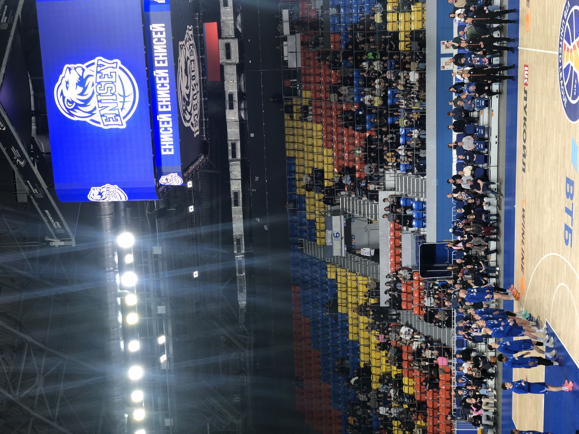 Here is a detailed description of the image:
The image shows the interior of a basketball arena, likely located in Krasnoyarsk, Russia, as indicated by the signage in the Cyrillic alphabet. The image captures a basketball court with the logo of VTB United League prominently displayed. A line of basketball players wearing blue uniforms are standing on the court. Seating rows are visible behind them, filled with spectators. The seats are colored in a pattern of blue, yellow, and red. Overhead, there is a large display screen with the logo of the team "Yenisey" prominently featured. The arena's lighting is bright, with multiple spotlights illuminating the scene.
