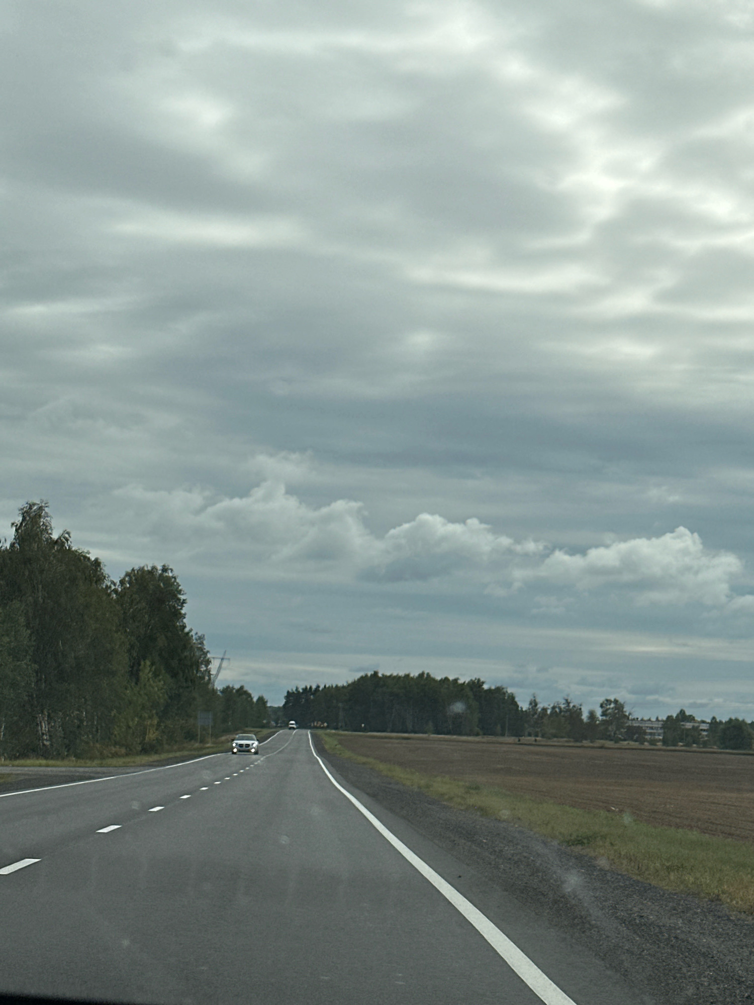 The image shows a view from a vehicle traveling on a two-lane asphalt road. The road has white dashed lines dividing the lanes and solid white lines on the outer edges. A dark green line of trees runs along the left side of the road, with some lighter green deciduous trees visible. To the right of the road is a large, plowed agricultural field with brown soil and some patches of green grass along the edge. In the distance, on the right side of the field, there are some buildings and more trees.
Two white cars are visible on the road in the distance, both traveling in the same direction as the viewer. The car in the foreground on the road appears to be a light-colored sedan, possibly white or silver. The sky is overcast with thick, gray clouds, but there are some brighter areas where sunlight is breaking through. The overall impression is of a rural landscape on a cloudy day. The location is specified as Paritsky Selsoviet, Belarus.
