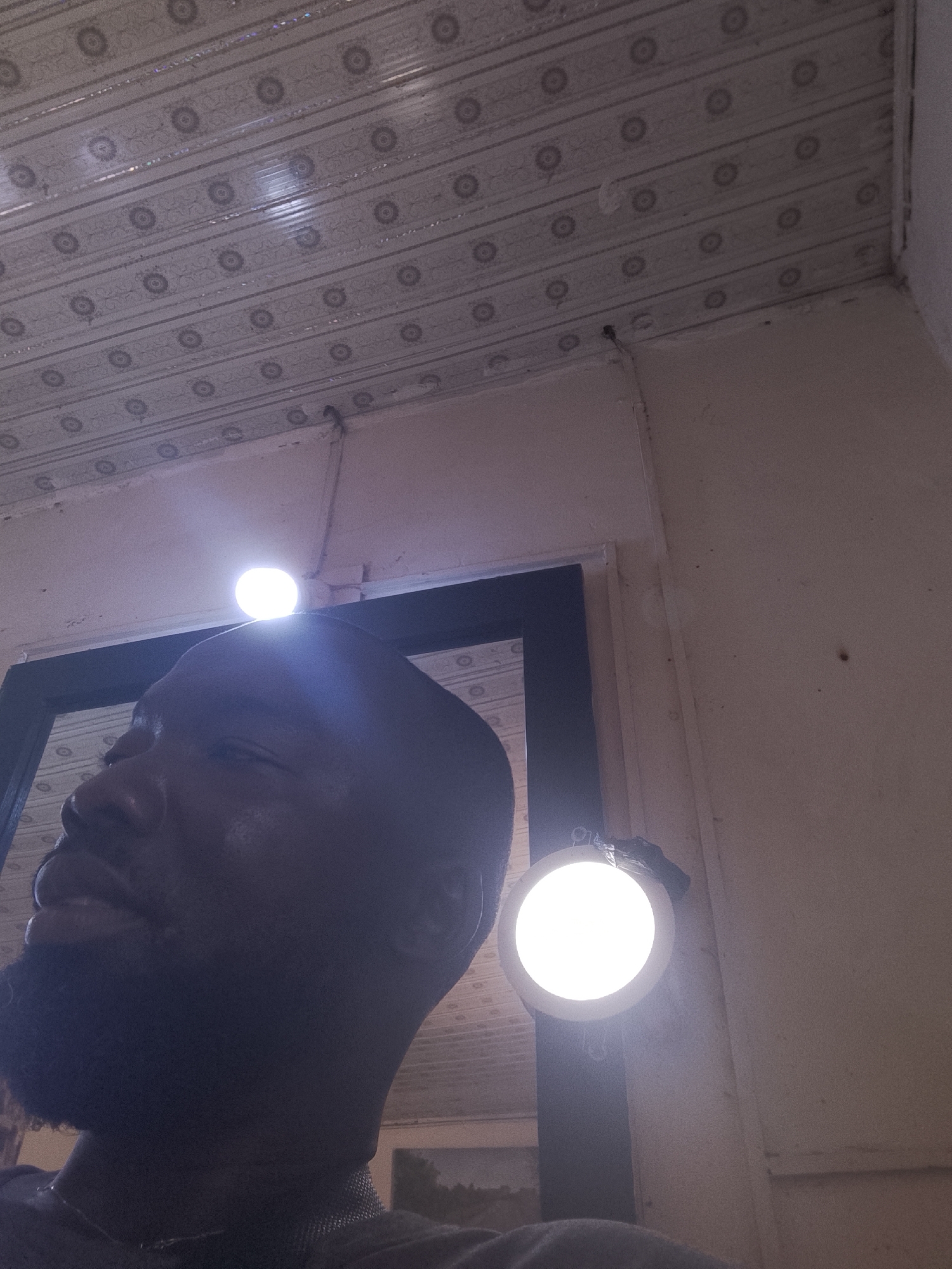 A low-angle shot depicts a dark-skinned individual with a beard, positioned between two illuminated circular lights affixed to a doorframe or wall. The ceiling above is textured with a patterned design. The setting appears to be an indoor space with light-colored walls. The city and specific location are unknown.