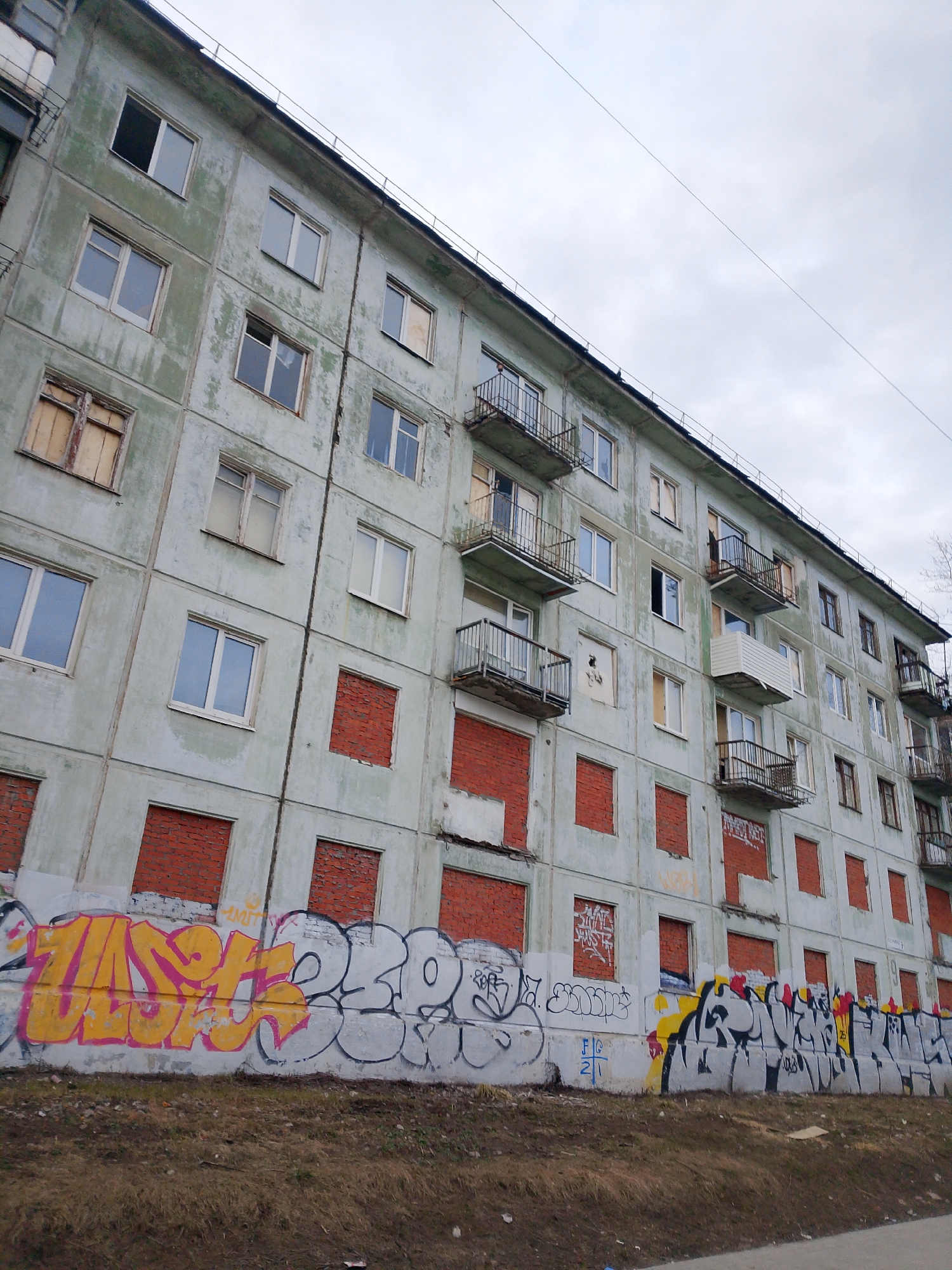 A low-angle shot of an apartment building in Izhevsk, Russia, reveals a weathered, grey facade covered in graffiti along the base. Many windows are bricked up or boarded over. Some windows are intact and reflect the cloudy sky. Numerous small balconies with metal railings extend from the building. The ground in front is covered with dirt and sparse vegetation. The sky is overcast and grey. A power line runs diagonally across the upper right portion of the image.