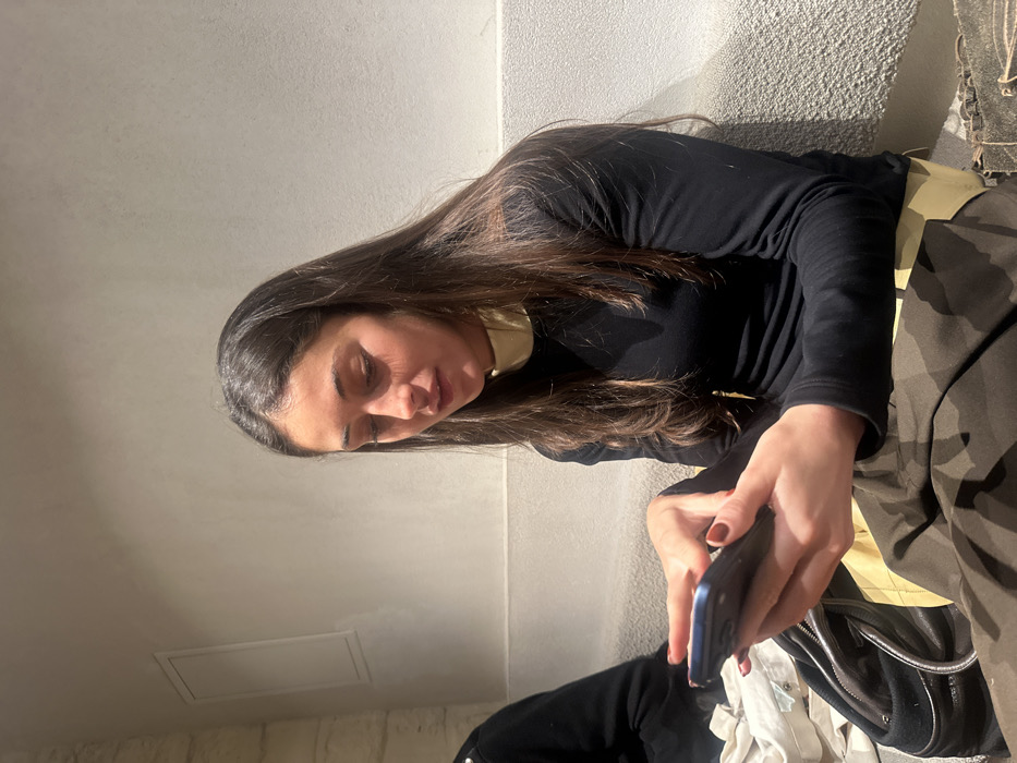 This is an indoor, vertically oriented photograph depicting a young woman absorbed in her smartphone. The subject, a woman with long, straight dark brown hair, is positioned in the center and right side of the frame, looking down at a device held in her hands. She appears to be in her 20s or 30s, with a focused or contemplative expression.
She is dressed in a black long-sleeved top, with a light-colored, possibly cream or beige, collar visible underneath. On her lower body, dark, possibly camouflage-patterned fabric is visible on the right side of the image. Her fingernails are painted a dark red.
The woman is holding a blue smartphone with both hands, positioned in front of her. To her right, a black bag with a metallic zipper is visible, along with some lighter-colored fabric underneath it, suggesting personal belongings.
The background consists of a light beige or off-white wall, which appears textured. A horizontal line suggests a seam or corner in the wall behind her. In the lower left portion of the image, a rectangular panel or vent is visible on the wall. The lighting is even, with soft shadows indicating an indoor setting, possibly lit by natural light from a window or well-distributed artificial light. The overall scene suggests a casual moment indoors, with the woman engrossed in her phone. The location context provided is Paris, France. No visible text is present in the image.