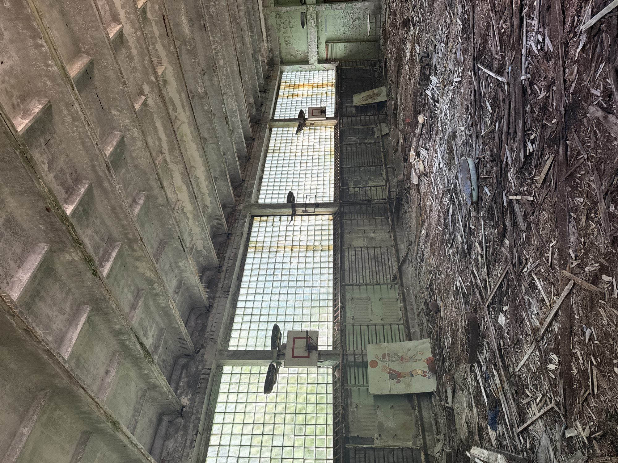The image depicts the interior of a derelict gymnasium in the city of Prypiat, Ukraine. The space is in a state of advanced decay, with a concrete ceiling featuring visible beams and water damage. Large, grimy glass block windows line the walls, allowing diffuse light to enter. The floor is heavily littered with debris, including broken wooden planks, scattered rubble, and what appears to be remnants of old equipment.
Prominently featured are two basketball hoops, mounted on poles, with faded backboards. To the left, leaning against the wall, is a large, faded poster or painted canvas depicting two figures playing basketball. In the foreground, there is a rusty metal grate on a circular base and other scattered wooden debris. The walls are painted in muted, peeling colors, predominantly a light green. The overall impression is one of abandonment and the passage of time, consistent with other structures in the exclusion zone of Prypiat. No people or discernible recent activity are present.
