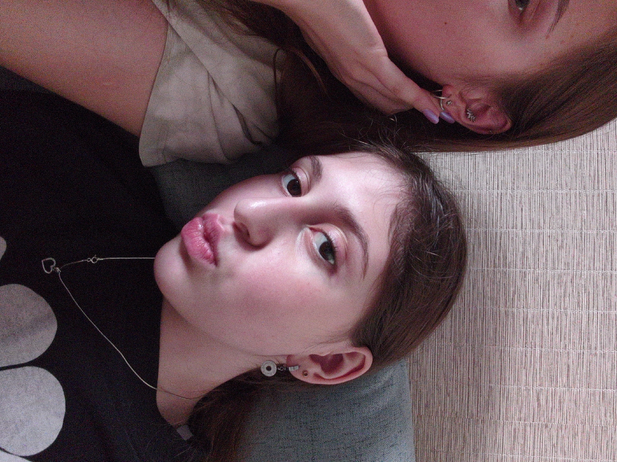 A selfie-style photograph shows two females indoors. One female is lying down, facing upwards, with a silver necklace and multiple earrings visible. The other female is partially visible in the upper right corner, with a manicured hand near her ear, adorned with multiple earrings as well. The location appears to be indoors, with a textured wall or mat in the background. No location in the city of Unknown, Unknown, can be inferred.