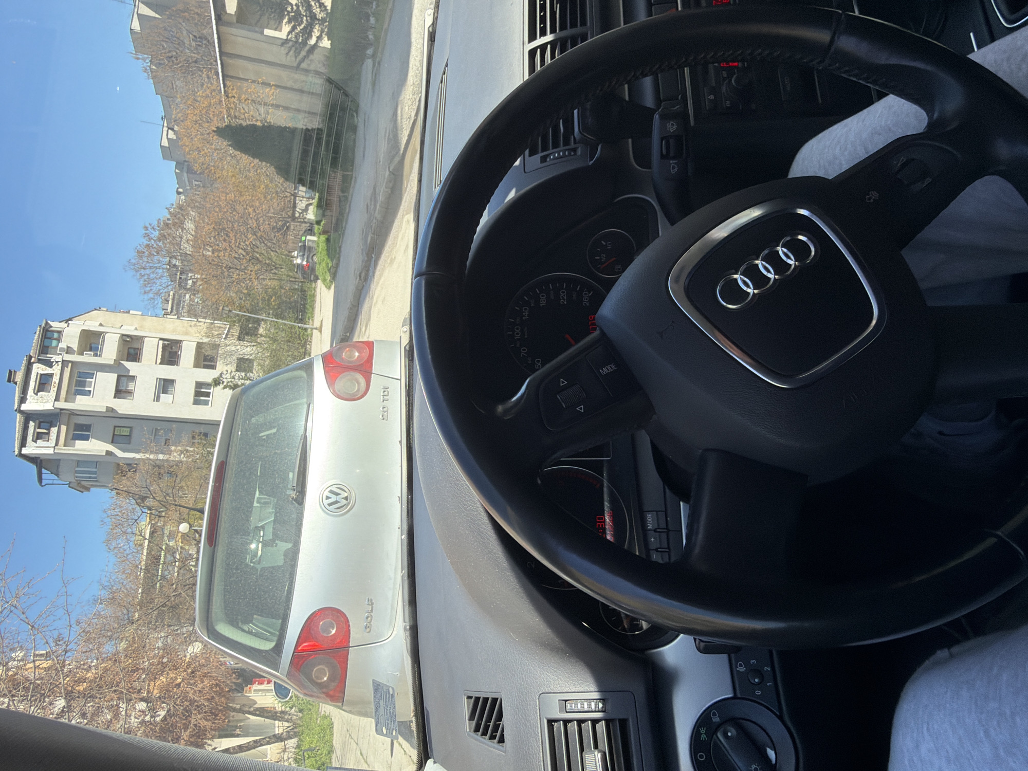 The image shows the interior of an Audi car, specifically the steering wheel with the Audi logo and parts of the dashboard. The view outside the front window reveals a silver Volkswagen Golf parked on the street. The surrounding environment includes residential buildings and trees typical of an urban setting. The text "2.0 TDI" is visible on the VW Golf, indicating it is a diesel model. The location is identified as Plovdiv, Bulgaria, based on additional contextual information. The time on the Audi's dashboard display shows "1:30". The driver is wearing a gray sweatshirt.