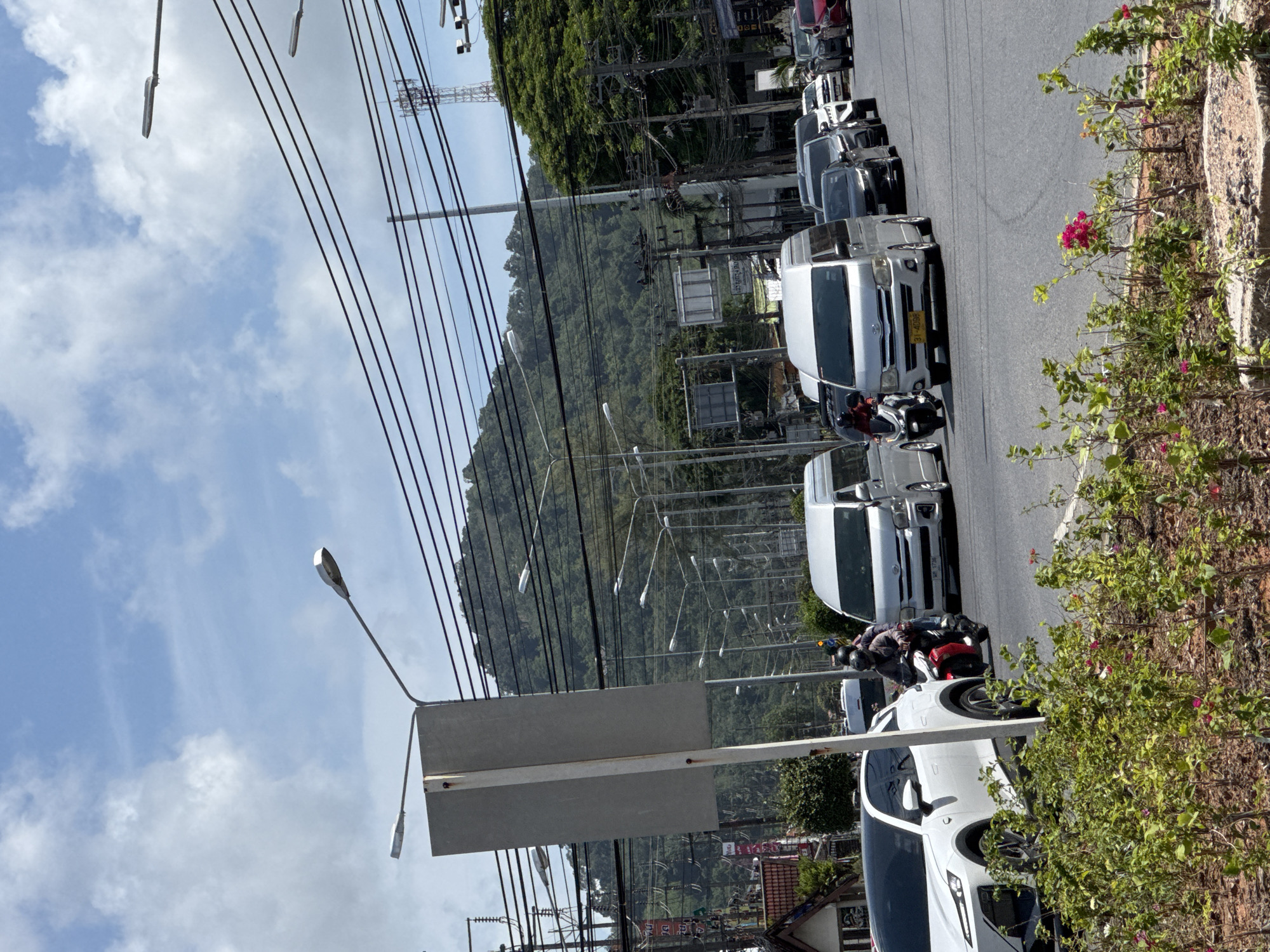 This image depicts a street scene in Kathu, Thailand, with a focus on traffic and the surrounding environment. In the foreground, a row of green bushes with pink flowers is visible, bordering the road. The road itself is paved and appears to be an intersection, with multiple lanes of vehicles.
Several cars and vans are present, including a white SUV with a black roof on the left, a silver minivan in the center, and other vehicles of various colors and types lined up behind it. A red motorcycle with two people wearing helmets is maneuvering through the traffic. Overhead, a complex network of power lines and streetlights crisscrosses the sky. In the background, a densely forested green mountain rises above the urban landscape. The sky is blue with scattered white clouds, indicating a daytime scene. The overall impression is one of a busy urban environment with a tropical backdrop.