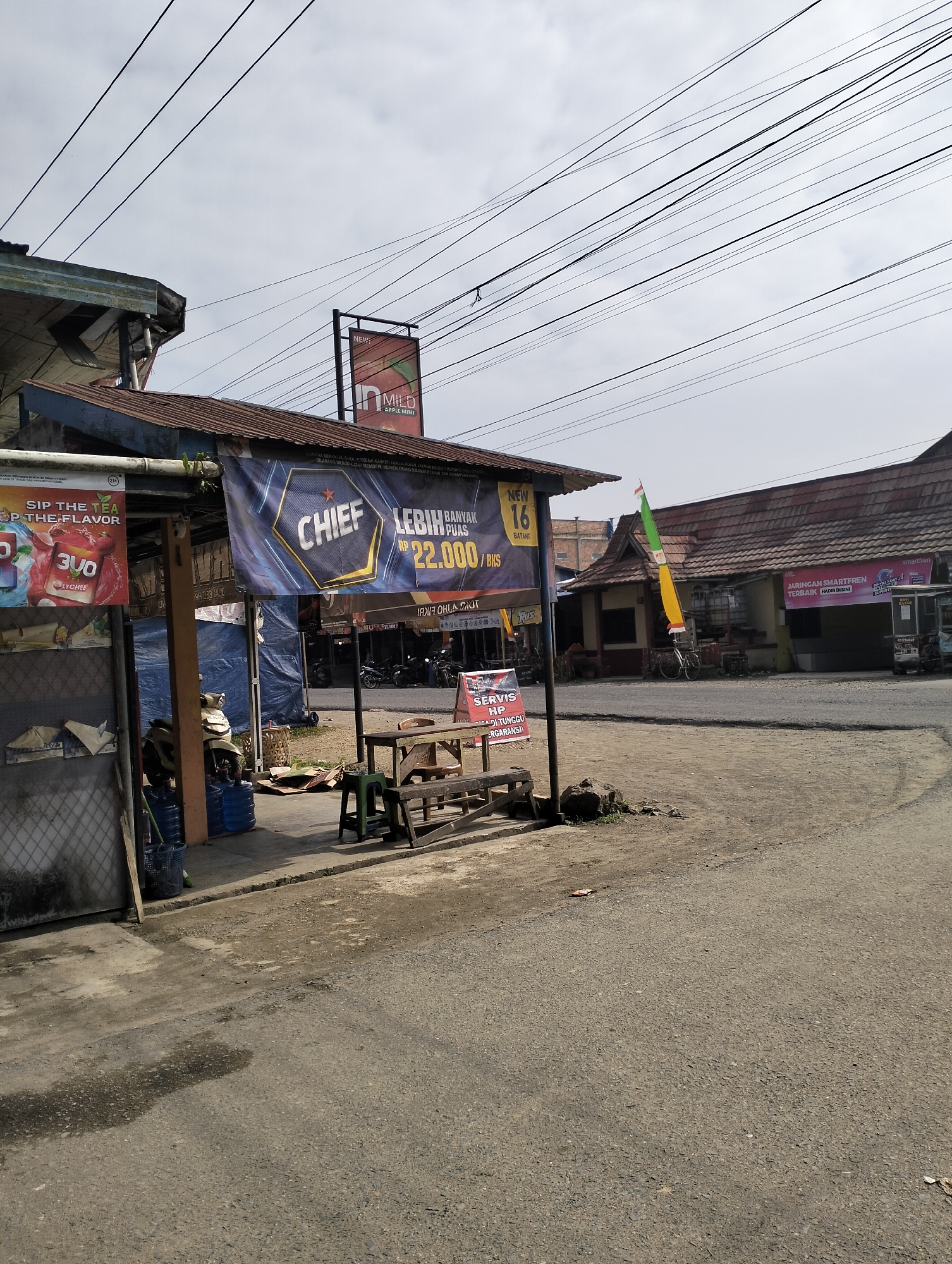 The image depicts a street scene in Pematang Lumut, Indonesia, likely a rural or semi-urban area, characterized by weathered buildings, overhead electrical wires, and a paved road. The foreground is dominated by a dirt and asphalt road with some puddles. To the left, a structure with a corrugated metal roof has a storefront with various advertisements. One banner prominently displays "CHIEF LEBIH BANYAK PUAS" with a price of "Rp 22.000 / BKS". Another banner on the left advertises "SIP THE TEA... THE FLAVOR" with imagery of a drink. Adjacent to the storefront, several large blue water jugs are visible, along with a black trash bin.
A simple wooden seating area with benches and a small table is positioned on the side of the road, suggesting a resting spot or a small roadside stall. Behind this, a building with a red-tiled roof and traditional Indonesian architectural elements is visible. A bicycle is leaning against the side of this building. Further in the background, more buildings and a few motorcycles are apparent, indicating some local commerce or residential activity. The sky is overcast. The overall impression is one of a modest, functional roadside environment in Indonesia.