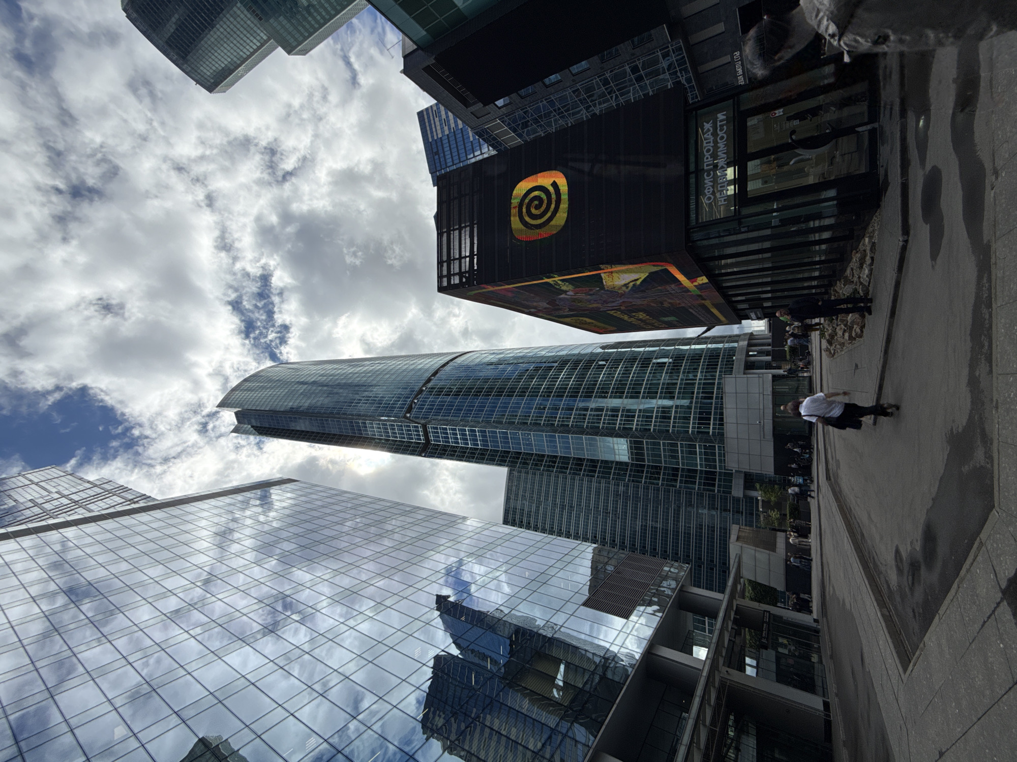 This is a low-angle, wide shot of a modern urban landscape, likely taken in Moscow, Russia, given the location cues. The scene is dominated by towering skyscrapers with reflective glass facades, interspersed with clouds in a dynamic sky. The buildings exhibit a sleek, contemporary architectural style.
In the foreground, a person on a skateboard or scooter is captured mid-action, their movement blurred due to the shutter speed, suggesting a daytime activity. Further in the background, other individuals are visible, walking along a paved pathway that borders the buildings. The ground appears wet in patches, hinting at recent rain or a damp environment.
One prominent building features a large, illuminated advertisement or display with a distinctive golden spiral logo against a black background. Below this, text in Russian is visible on a storefront or building facade, reading "ОФИС ПРОДАЖ НЕДВИЖИМОСТИ" (OFFICE FOR REAL ESTATE SALES).
The lighting suggests it is daytime, with the sun likely partially obscured by clouds, creating a diffused light that enhances the reflections on the glass buildings. The overall atmosphere is one of a bustling, modern city center with active pedestrian and possibly recreational movement.