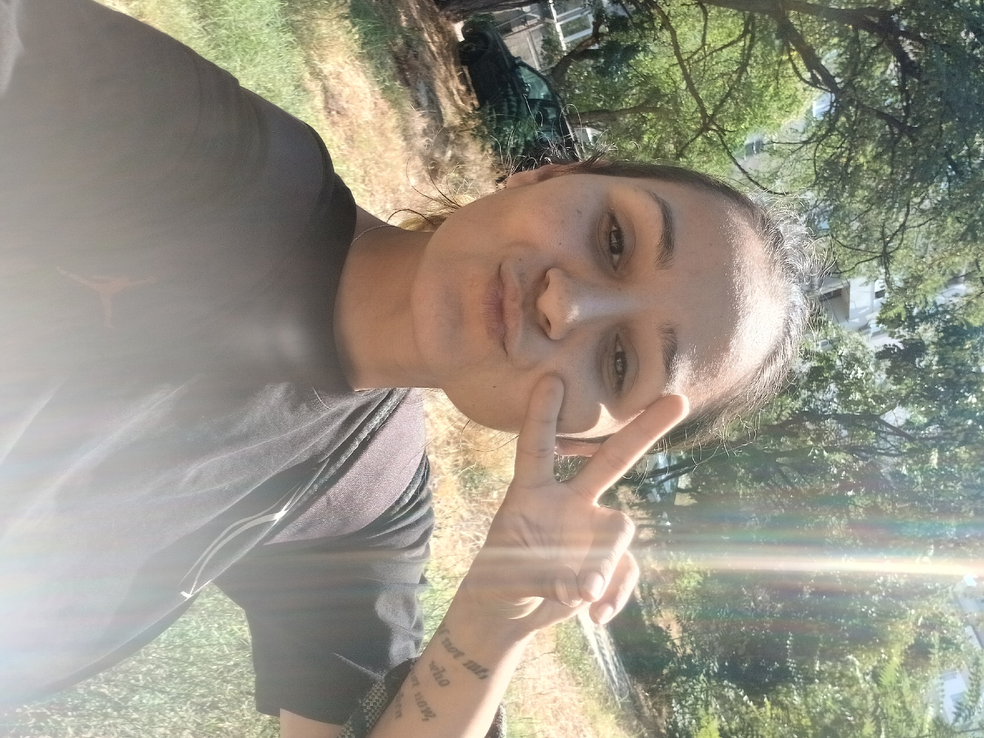 A young woman with dark hair pulled back into a bun takes a selfie outdoors. She is wearing a black t-shirt. Her right hand is raised in a peace sign, with her index and middle fingers extended. She has a tattoo on her left forearm with text that is partially visible. She is making a kissing face. In the background, there are trees, dry grass, a car, and a building. The image appears to have been taken on a sunny day, with lens flare from the sun. The location is not discernible from the image.