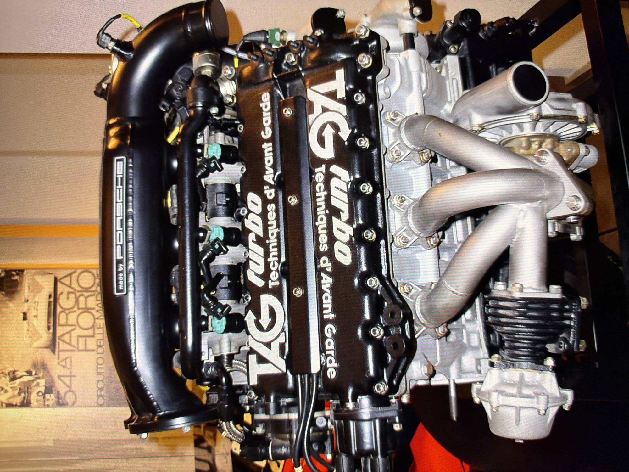 The image displays a close-up, eye-level view of a black Porsche engine, prominently featuring the "TAG Turbo" branding on its valve covers. The engine is the primary subject, with intricate details of its components visible, including silver exhaust manifolds, various hoses, and electrical connectors. The background features a poster with text that appears to be in Italian, possibly related to a racing event. The overall setting suggests an indoor display, perhaps in a museum or showroom. There are no people or animals present in the image, nor are there any indications of the city of Kyiv, Ukraine.