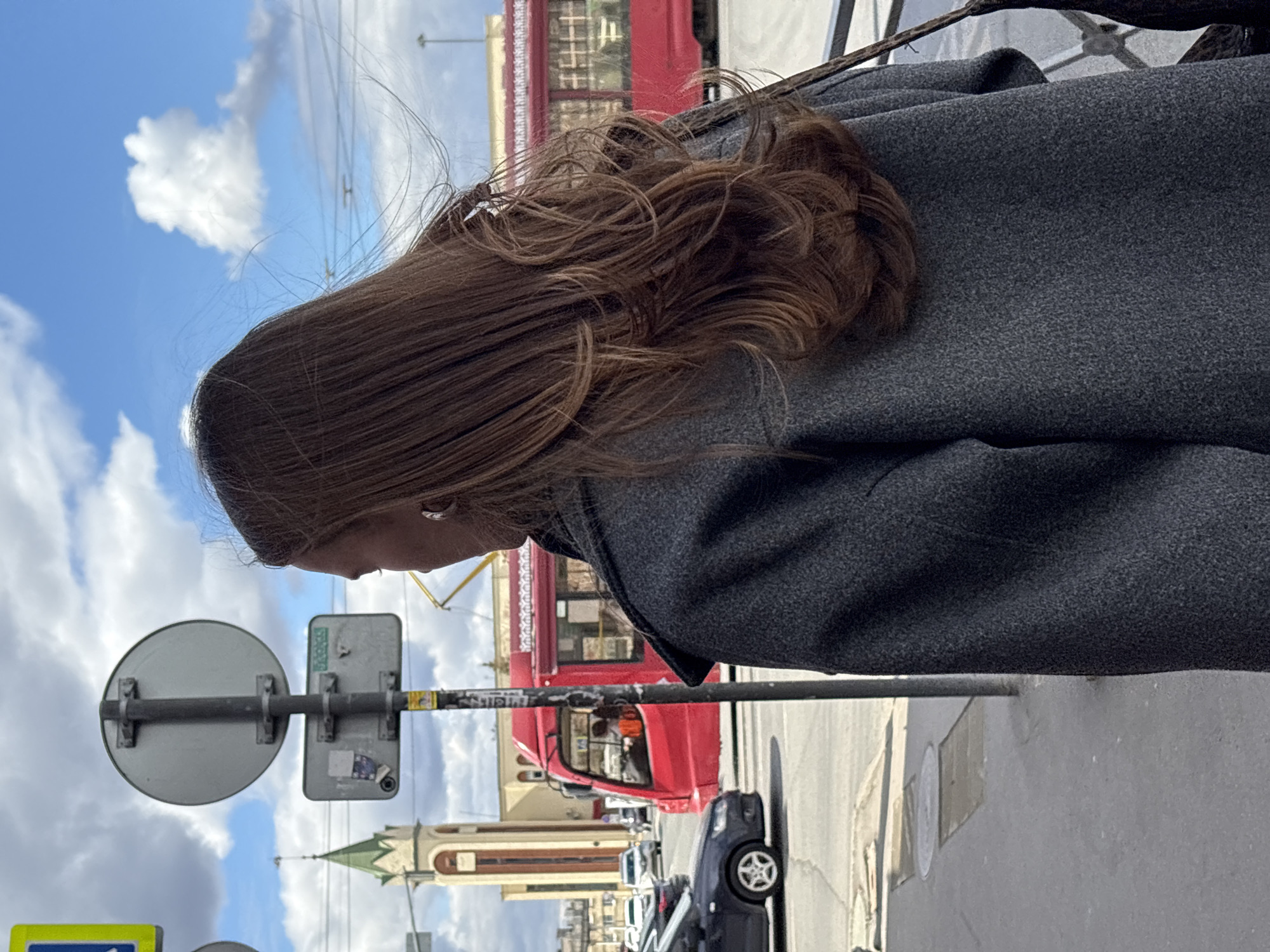 A medium, vertical shot shows a woman with brown hair wearing a dark gray coat standing on a street in Kazan, Russia. The woman is facing away from the camera and towards the street. A red bus is partially visible in the background, along with several parked cars. A street sign is in the foreground, obscuring part of the bus and the street. The sky is blue with some clouds. A church steeple is visible in the background.