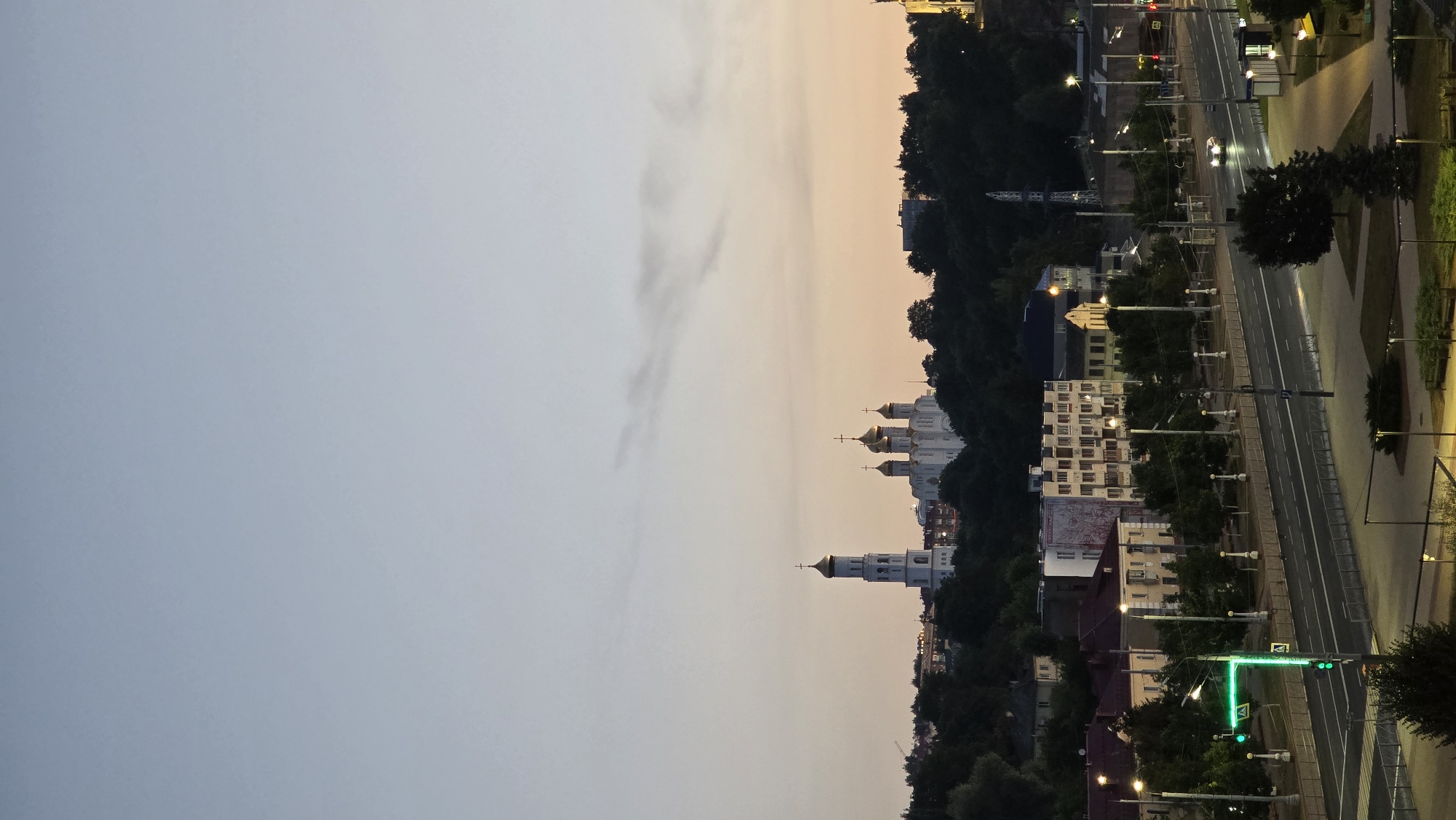 The image captures a twilight view of the city of Bryansk, Russia, with a prominent white cathedral topped with golden domes situated on a hill in the background. To the left of the cathedral, a tall bell tower, also capped with gold, stands out. The foreground features a multi-lane road with traffic lights, streetlights, and parked cars. Several buildings, including residential structures and businesses, line the street. Trees are abundant throughout the scene, creating a lush, green landscape that contrasts with the urban elements. The sky exhibits a gradient of pale blue and soft orange, indicating the transition from day to night.