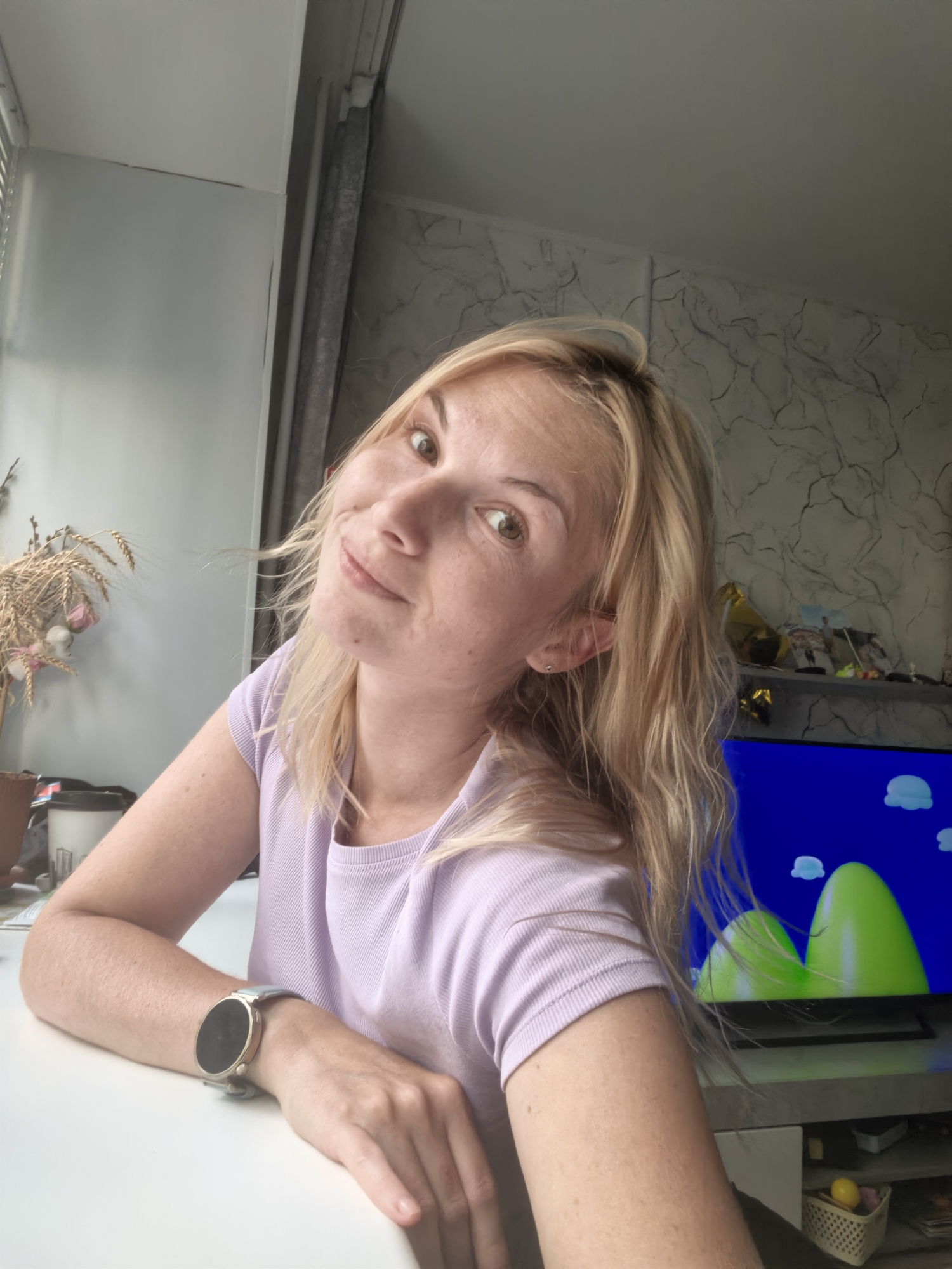 A woman with blonde hair and fair skin is looking at the camera, tilting her head slightly. She is wearing a light purple t-shirt and a smartwatch on her left wrist. She is sitting at a white table, with her left arm resting on it. In the background, there is a window with blinds on the left, a decorative plant on the table, and a television displaying a cartoon image of green hills and blue sky on the right. The wall behind the television has a textured, marble-like pattern. There are shelves above the television with various items. The overall scene appears to be an indoor setting, possibly a living room or office, in Sterlitamak, Russia.