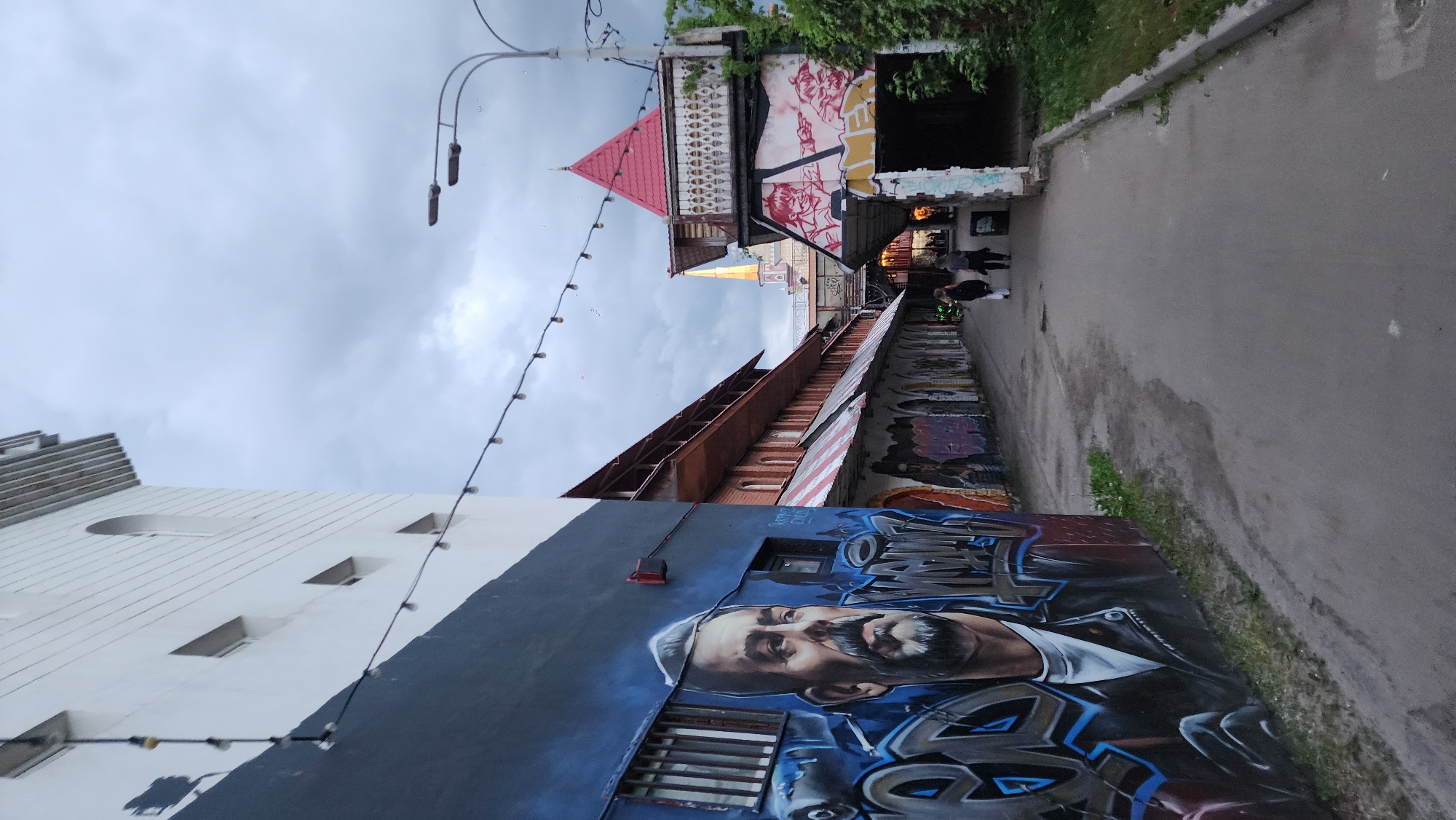 The image depicts a narrow street scene, possibly in Moscow, Russia, characterized by colorful street art and architectural diversity. The walls lining the street are covered in graffiti, including a large mural of a man with a beard. Architectural styles vary, with buildings displaying red roofs and decorative elements. The sky is overcast. Two people are walking down the street. Overhead, string lights stretch across the street.