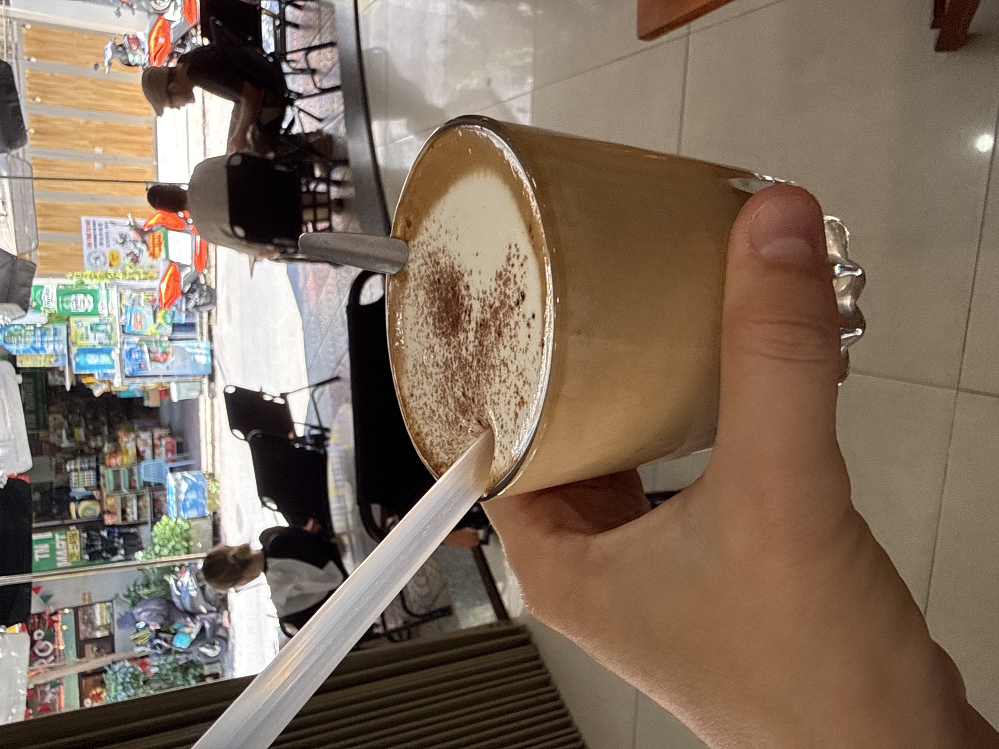 A hand holds a glass of cappuccino with a straw in an indoor setting. The cappuccino has a layer of white foam on top, dusted with brown powder. Outside the window, the reflection shows a view of a street in Đà Nẵng, Vietnam. A shop displays merchandise, and people are seated at tables, suggesting a café or restaurant. A street with parked scooters and motorbikes is visible.