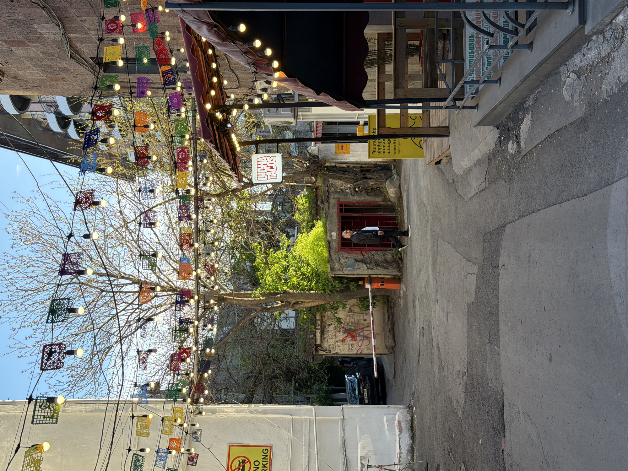 The image captures a vibrant, decorated alleyway in Yerevan, Armenia, during the daytime under a clear blue sky. Overhead, strings of lights are interwoven with colorful, cut-paper banners (papel picado style), creating a festive atmosphere. Bare tree branches, characteristic of early spring, are visible against the sky on the left.
On the right side of the alley, a man wearing a dark jacket and light shirt stands near a building entrance with a red frame, appearing to observe the scene. Business signs are prominent: a white sign for "TACOS" is visible, along with a yellow sign displaying "Yandex Ea" and Armenian text, which includes phrases like "Պատվիրեք սիրելի ուտեստ" (Order favorite food) and "առաք" (delivery). A "NO PARKING" sign with a red prohibition symbol is attached to a building on the left.
The ground is a mix of asphalt and concrete. Further down the alley, a red and white barrier arm is present, and a black car with the license plate "70 OX 007" is partially visible. A wall behind the barrier shows graffiti. The overall scene suggests a lively urban commercial area, likely featuring restaurants or cafes, enjoying pleasant, bright weather.