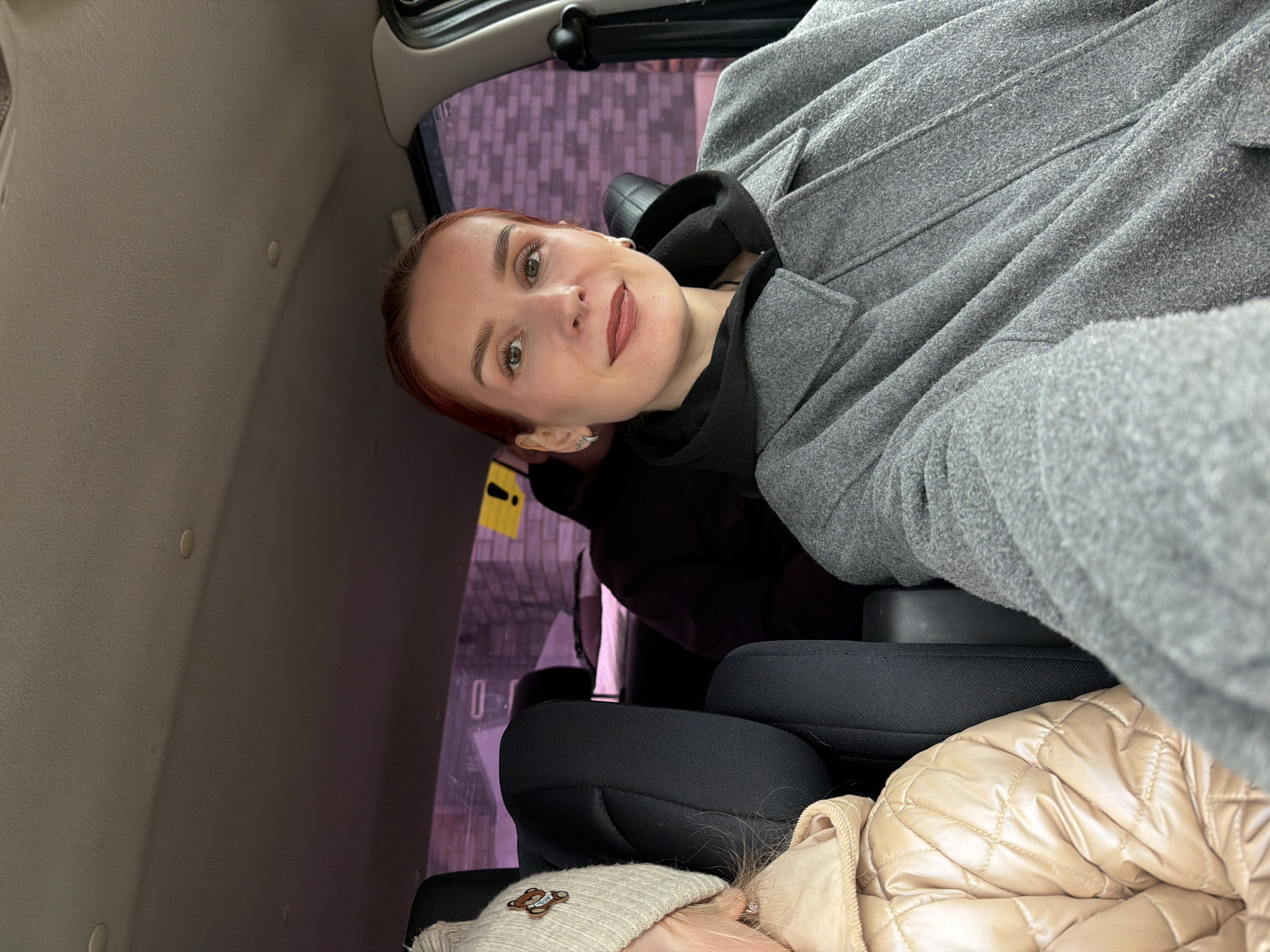 The image is taken inside a car, possibly in Chaikovsky, Russia. A person with reddish-brown hair is looking at the camera while wearing a gray coat and a black hoodie underneath. The person is sitting in the back seat of the car, next to a car seat. Part of another person wearing a tan-colored puffy jacket and a beanie is also visible. The car's interior includes gray ceiling panels and window pillars. A glimpse of buildings can be seen through the car windows.