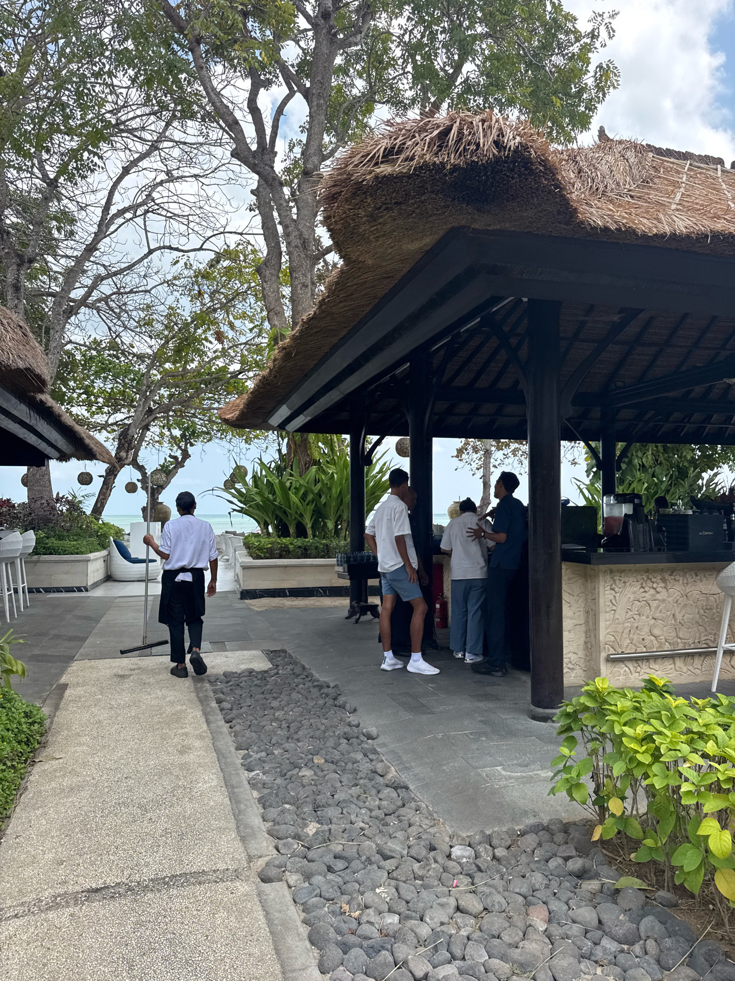 A restaurant with a thatched roof and open sides is situated near the ocean in Kuta Selatan, Indonesia. A waiter wearing a white shirt and black apron walks away from the camera carrying a pole. Several patrons are gathered at the bar inside the restaurant, interacting with bar staff. Lush green plants and trees surround the outdoor seating area, and the ocean is visible in the background. The ground is paved with stones and concrete.