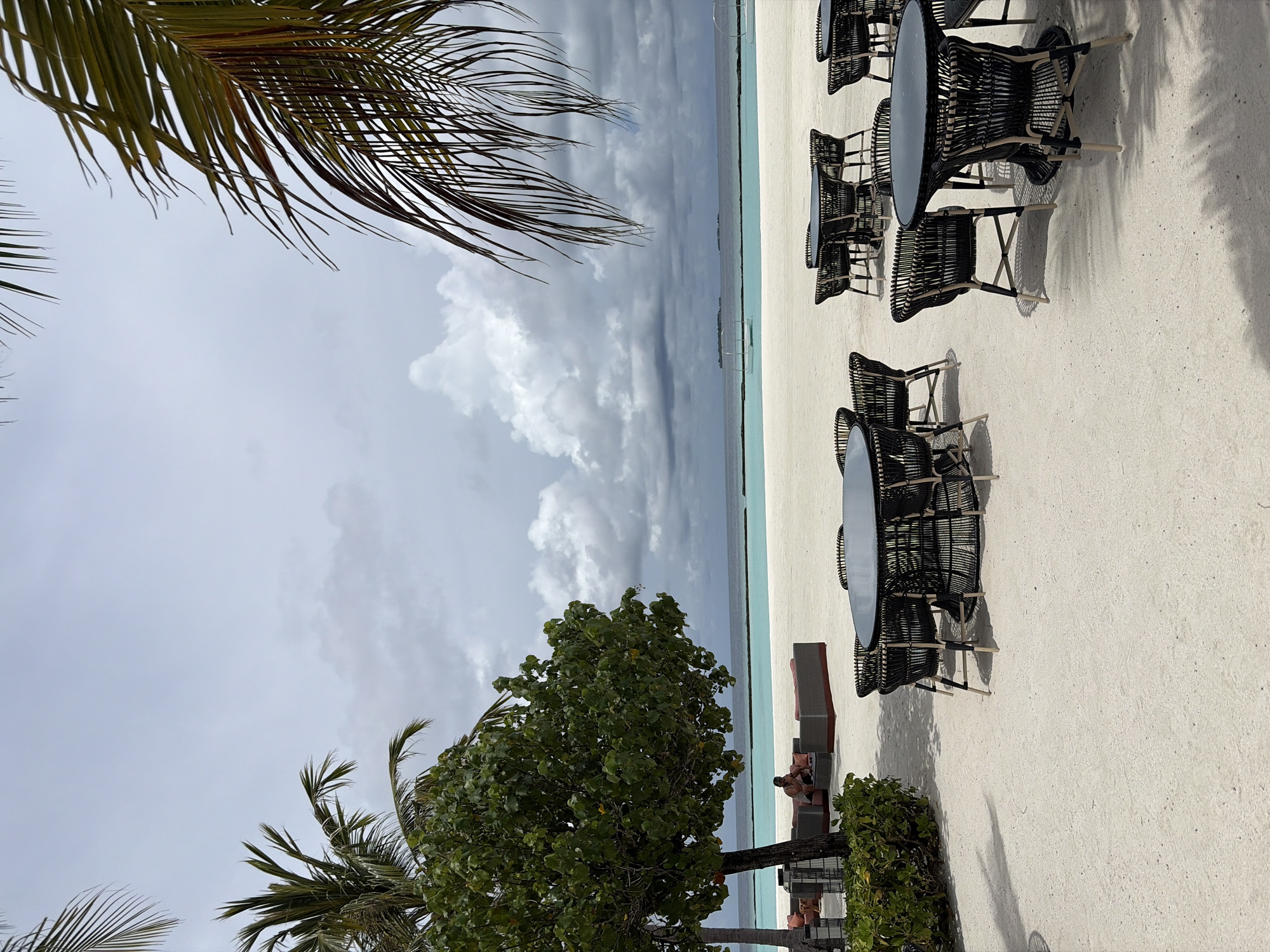 This is a photograph of a resort dining area on a sandy beach in Halaveli, Maldives. In the foreground, several black wicker tables and chairs are arranged on the white sand. Some of the tables have black tops and the chairs have light-colored legs. Palm fronds hang into the frame from the top right and top left corners. In the midground, a large green tree shades a seating area with orange cushioned couches. A single person is seated on one of the couches. Beyond the seating area is a turquoise ocean and a cloudy sky. The horizon line is clearly visible, separating the water from the sky.