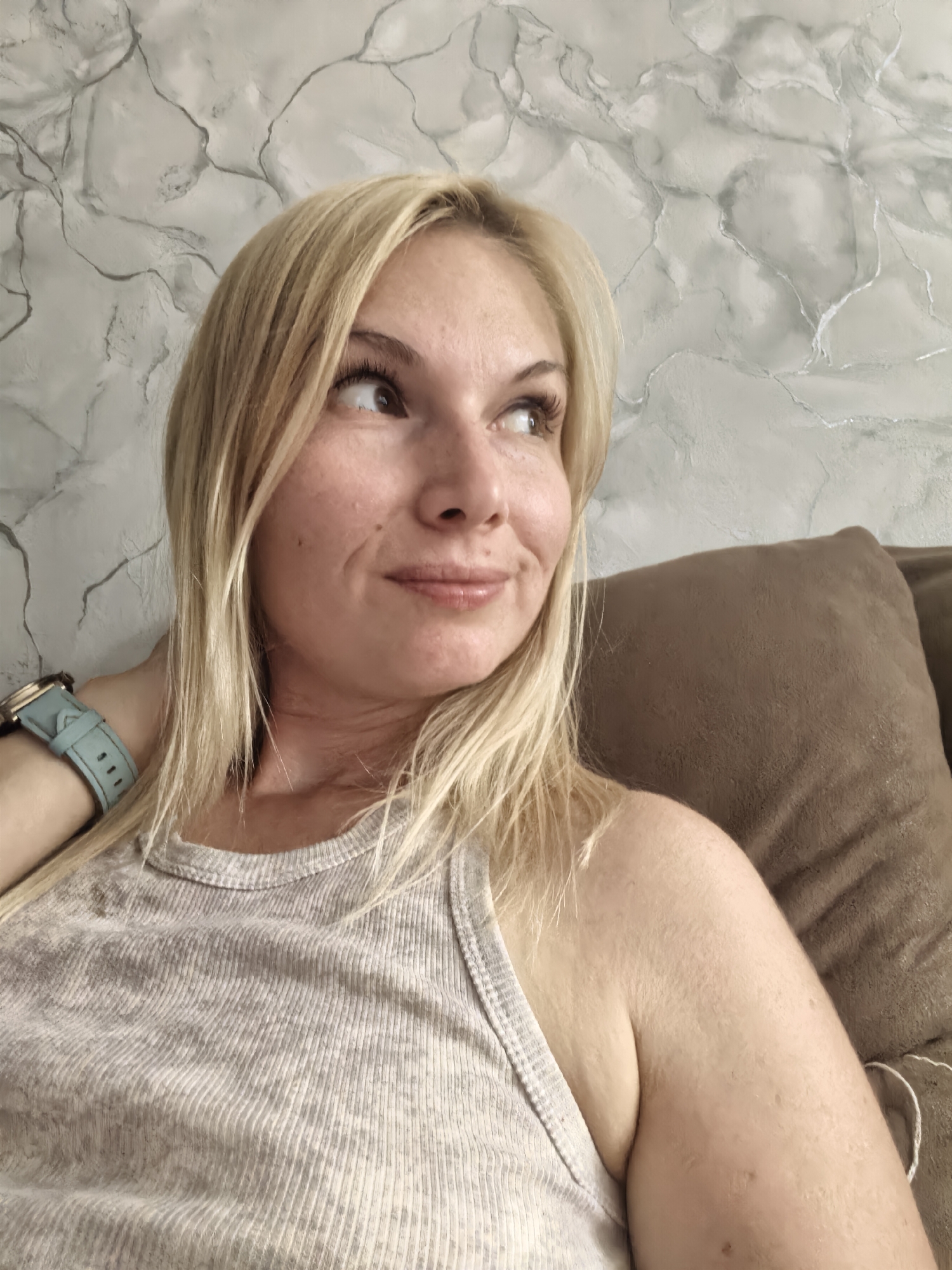 A medium shot shows a fair-skinned blonde woman from the chest up, looking to her right with a slight smile. She wears a light gray ribbed tank top and a light blue watch on her left wrist. Her blonde hair is shoulder-length and styled straight. The background is a textured wall with a marble-like pattern and a brown couch cushion on the right. The image appears to be taken indoors.