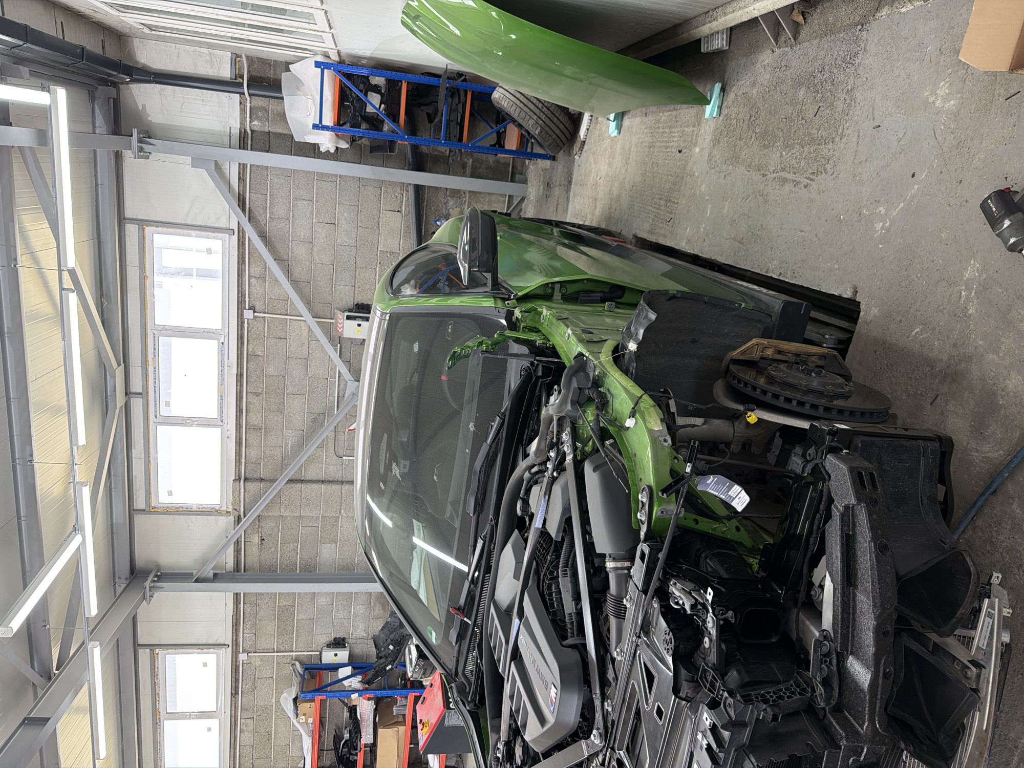 An overhead view captures a brightly lit indoor automotive workshop. The central subject is a heavily damaged bright green car, identified as a Toyota GR Supra (A90 generation) due to the visible "BMW M POWER" engine cover and a "GR" badge on a black plastic component near the engine bay. The car's front end is extensively disassembled and damaged, with the bumper, grille, and other body panels removed, exposing the engine, radiator, and underlying structural components. The front right wheel assembly, including the brake rotor and caliper, is visible without a tire. A separate green body panel, likely a fender or hood, leans against a concrete block wall in the background.
The workshop features concrete block walls and a concrete floor. In the background, blue and orange metal shelving units hold various car parts, including a tire and plastic components. Fluorescent strip lights are mounted on the ceiling structure, providing bright illumination. Windows are visible higher up on the walls, admitting natural light. On the floor, a black impact wrench, possibly a WURTH brand, is discernible. The overall scene suggests a vehicle undergoing significant repair or reconstruction.