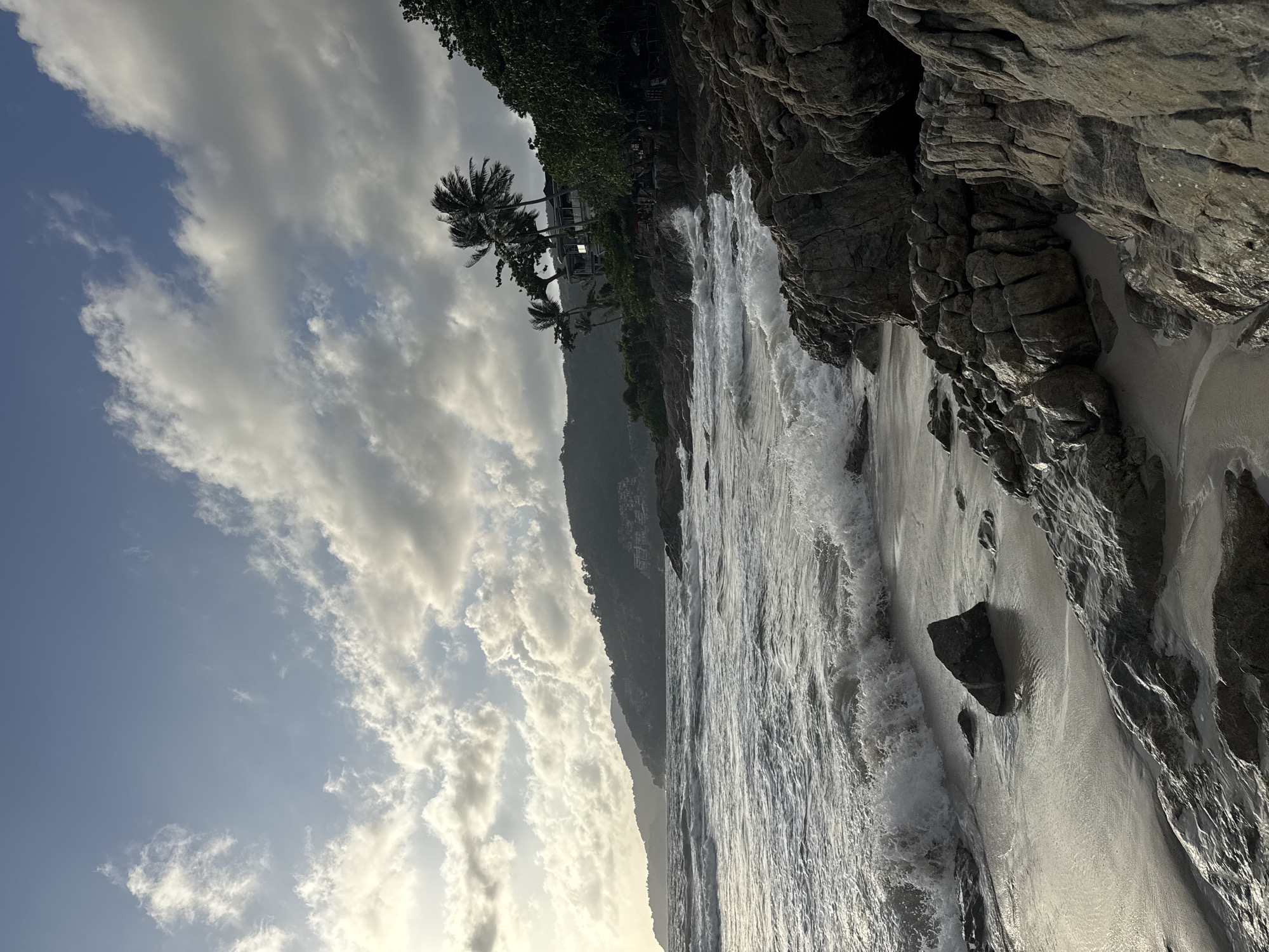 This image depicts a rocky beach in Patong, Thailand, during a cloudy day. The ocean waves are crashing onto the shore, creating white foam against the sand and rocks. The foreground is dominated by large, grey rocks with sandy patches between them. In the mid-ground, the sea stretches towards the horizon, with a line of green hills in the distance. Palm trees and a building are visible on the hillside, suggesting a resort or residential area. The sky is filled with scattered clouds, with patches of blue visible. The overall mood is natural and serene, with the raw beauty of the coastline on display.