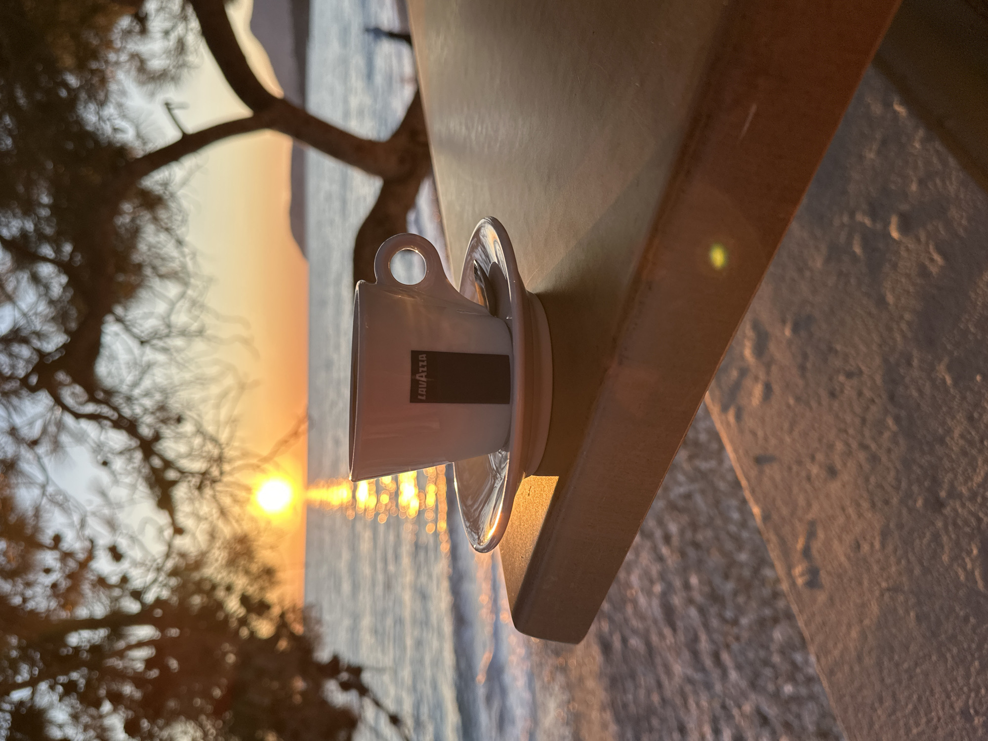 The image depicts a Lavazza coffee cup and saucer, positioned on a weathered, light-colored surface. The cup is white with a black Lavazza logo. The scene overlooks a shoreline at sunrise, with the sun reflecting off the water. Silhouetted trees frame the left side of the image. The location is the city of Bar, Montenegro. The foreground includes a textured wall-like surface. The scene is static and no people are visible.