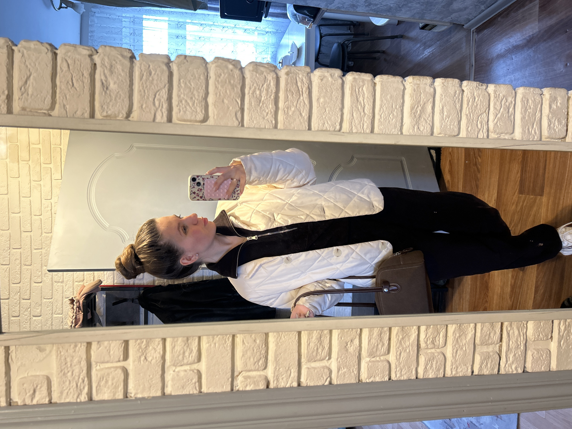 A woman is reflected in a mirror with a faux brick frame. She is holding a phone in her hand. She is wearing a black shirt, a white jacket, and black pants. She also has a brown purse. The reflection shows a wood floor and various items in the background, including clothing racks and furniture. The city of Klin, Russia is not discernible from the image.