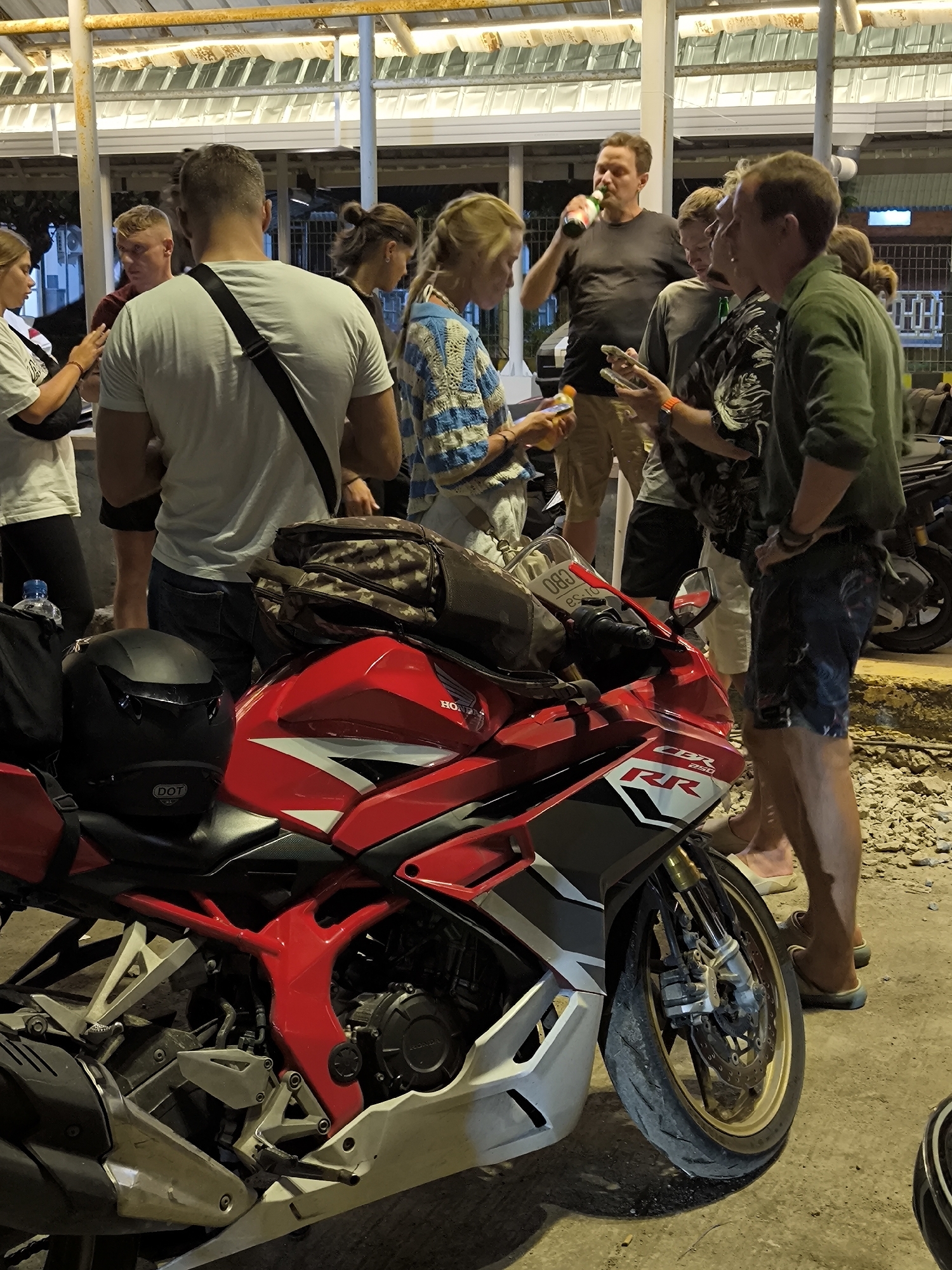 The image shows a group of about nine people standing and interacting around a red and white Honda CBR 250 RR motorcycle parked on a paved surface. A black helmet rests on the motorcycle's seat, and a camo bag is strapped on top of the motorcycle. The individuals appear to be tourists.
One man is drinking from a beer bottle. Several people are looking at their phones.
The background features a covered outdoor structure with visible support beams and a tiled roof. The location is likely a tourist gathering point, possibly a restaurant. Another motorcycle is parked in the background. The location, Unknown, Unknown, suggests that the city is not known. The time is likely evening or night.