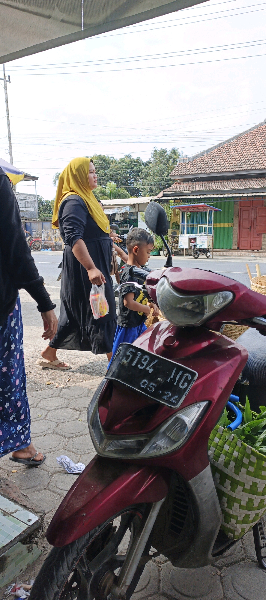 In Kasiyan, Indonesia, a woman wearing a yellow hijab and a black dress walks on a paved sidewalk, holding a clear plastic bag with yellow and red items inside. Beside her, a young boy in a black t-shirt with a yellow pattern and a blue skirt walks. In the foreground, a maroon scooter is parked. The scooter has a license plate that reads "P 5194 MG 05.24". A green and white woven basket containing leafy green vegetables is attached to the side of the scooter. In the background, there are buildings with tiled roofs, trees, and a food stall. The weather appears to be sunny with a few clouds.