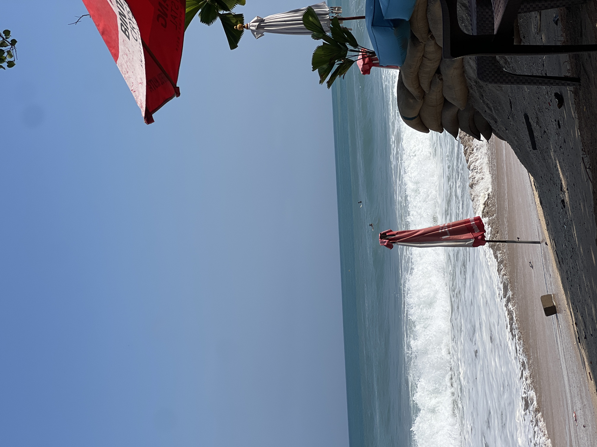 A serene beach scene unfolds under a clear blue sky in Kuta, Indonesia. The turquoise ocean meets a sandy shore with gentle waves rolling in, leaving a foamy white trail. In the foreground, a red and white beach umbrella, partially folded, stands erect on a metal pole, casting a shadow on the sand. To the right, a wall constructed from stacked sandbags rises from the beach, providing a protective barrier. A dark plastic chair sits at a table near the sandbags, suggesting a seating area for beachgoers. The background features lush green palm leaves and another tall, white umbrella. In the distance, a few surfers are visible in the water, enjoying the ocean's swell. The overall ambiance is peaceful and inviting, characteristic of a tropical beach destination.