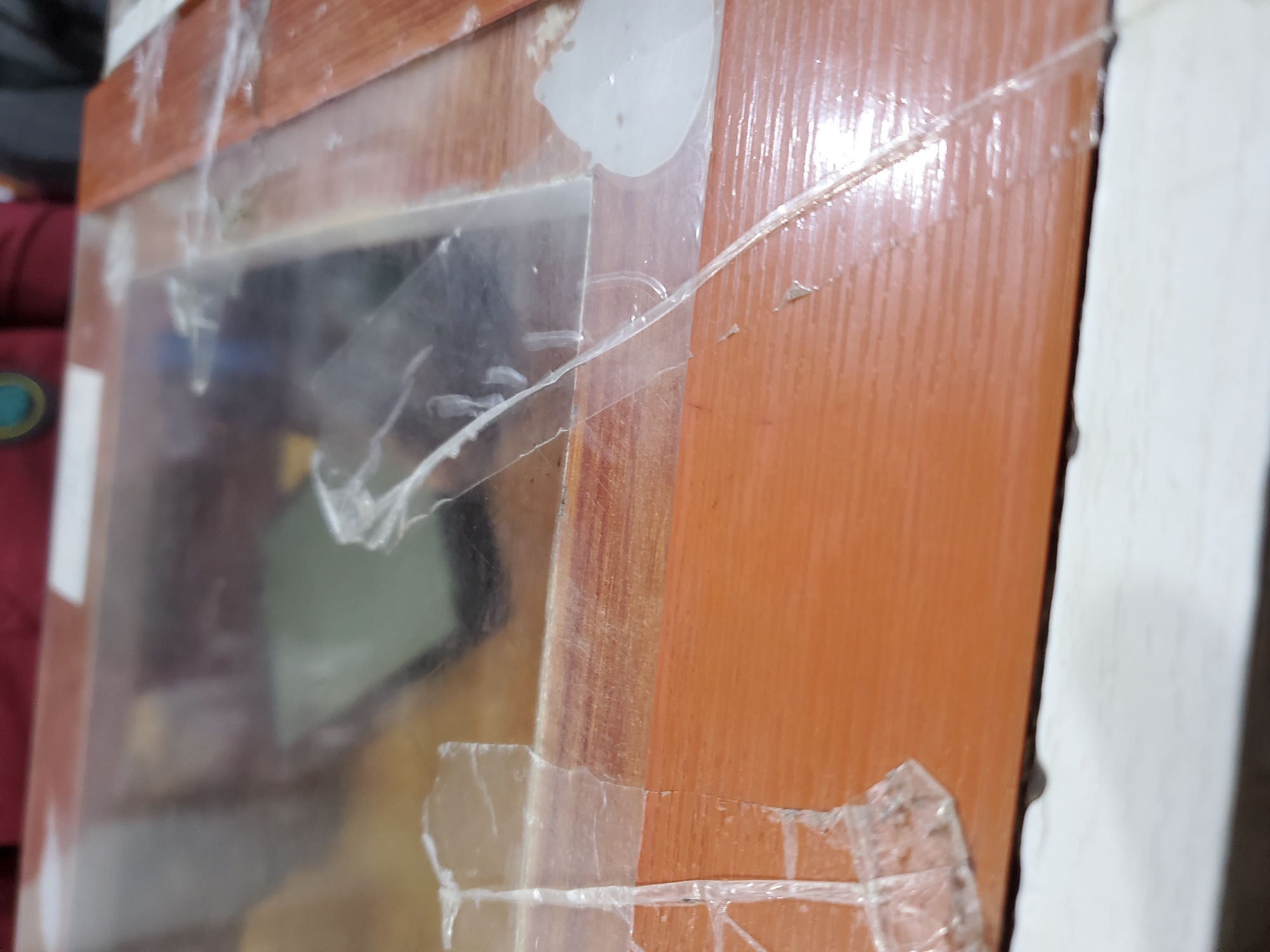 The image shows a close-up of a wooden surface, possibly a table or frame, with clear plastic sheeting taped over it. The wood has a reddish-brown, grainy texture. The plastic is creased, wrinkled, and has several pieces of clear tape holding it down. Some of the tape is peeling away, and there are smudges and distortions visible on the plastic surface. In the background, through the plastic, a dark object, possibly a phone, is visible, along with a blurred glimpse of fabric. The overall impression is one of a makeshift or temporary covering. There is no specific indication of the city of Jos, Nigeria, in the image.